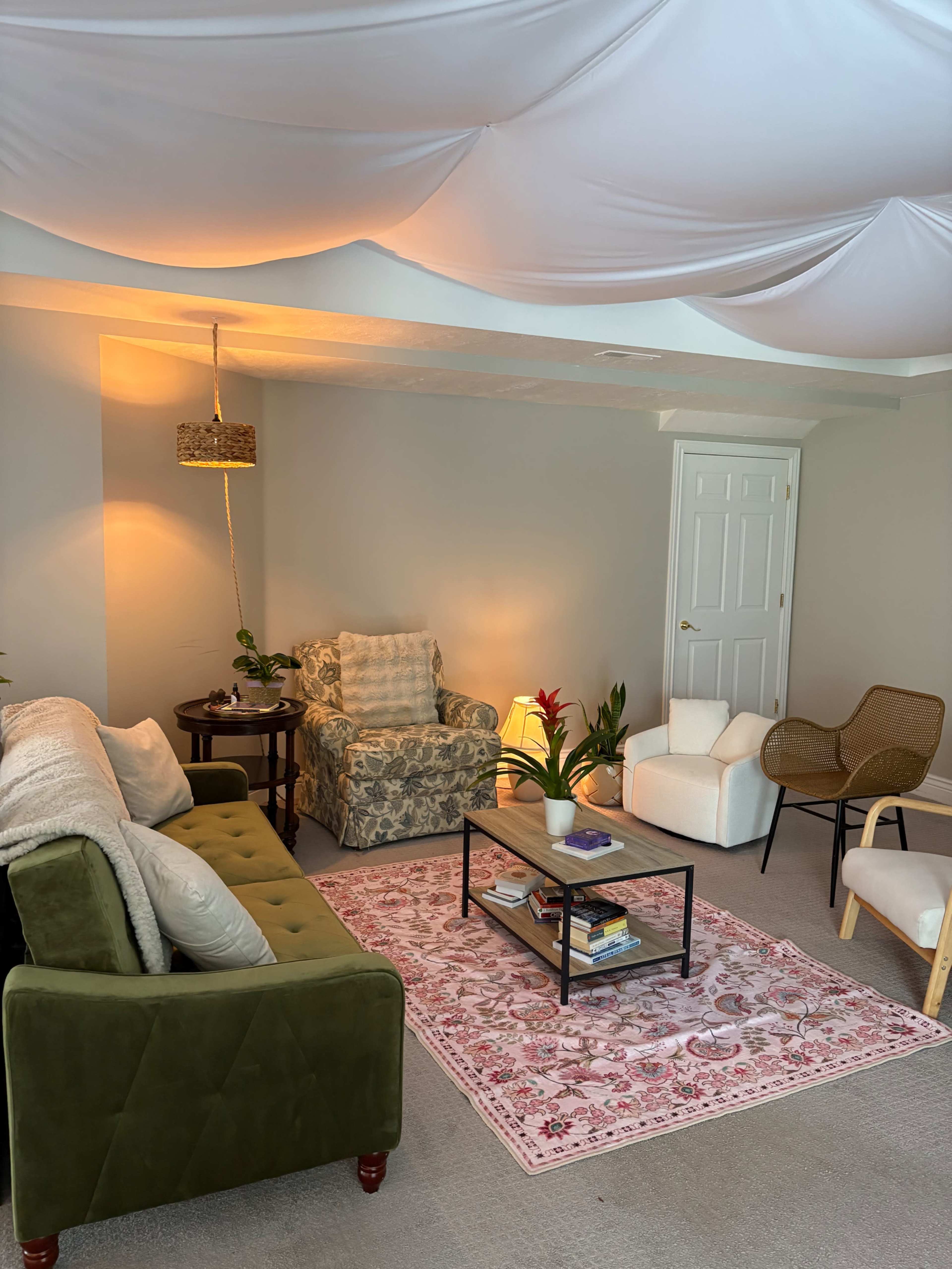 The image shows a cozy, well-decorated living space with a green velvet sofa, various chairs, a coffee table with books, and warm lighting under a draped ceiling.