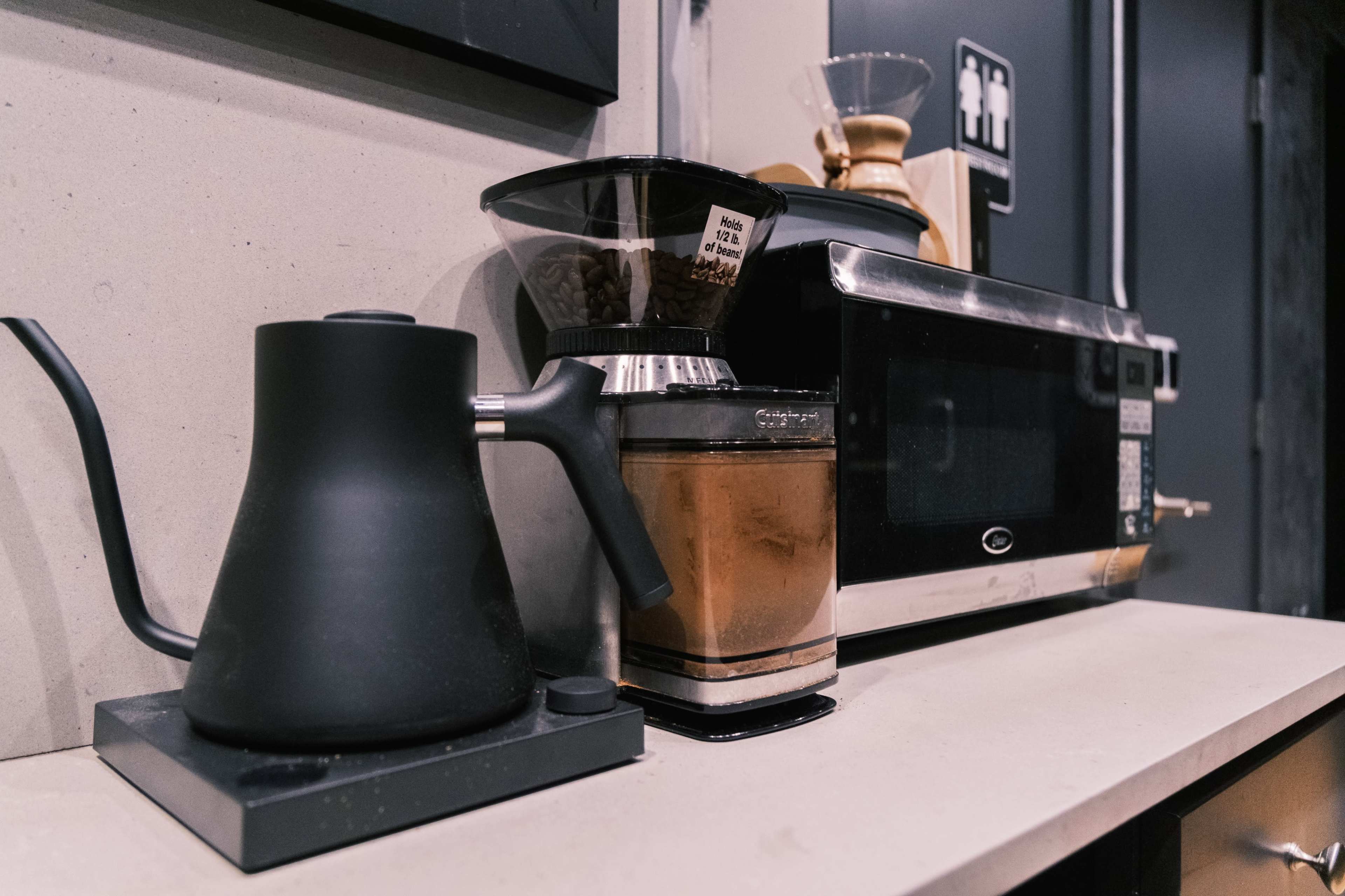 A black kettle and coffee grinder are placed on a countertop beside a microwave in a kitchen setting.