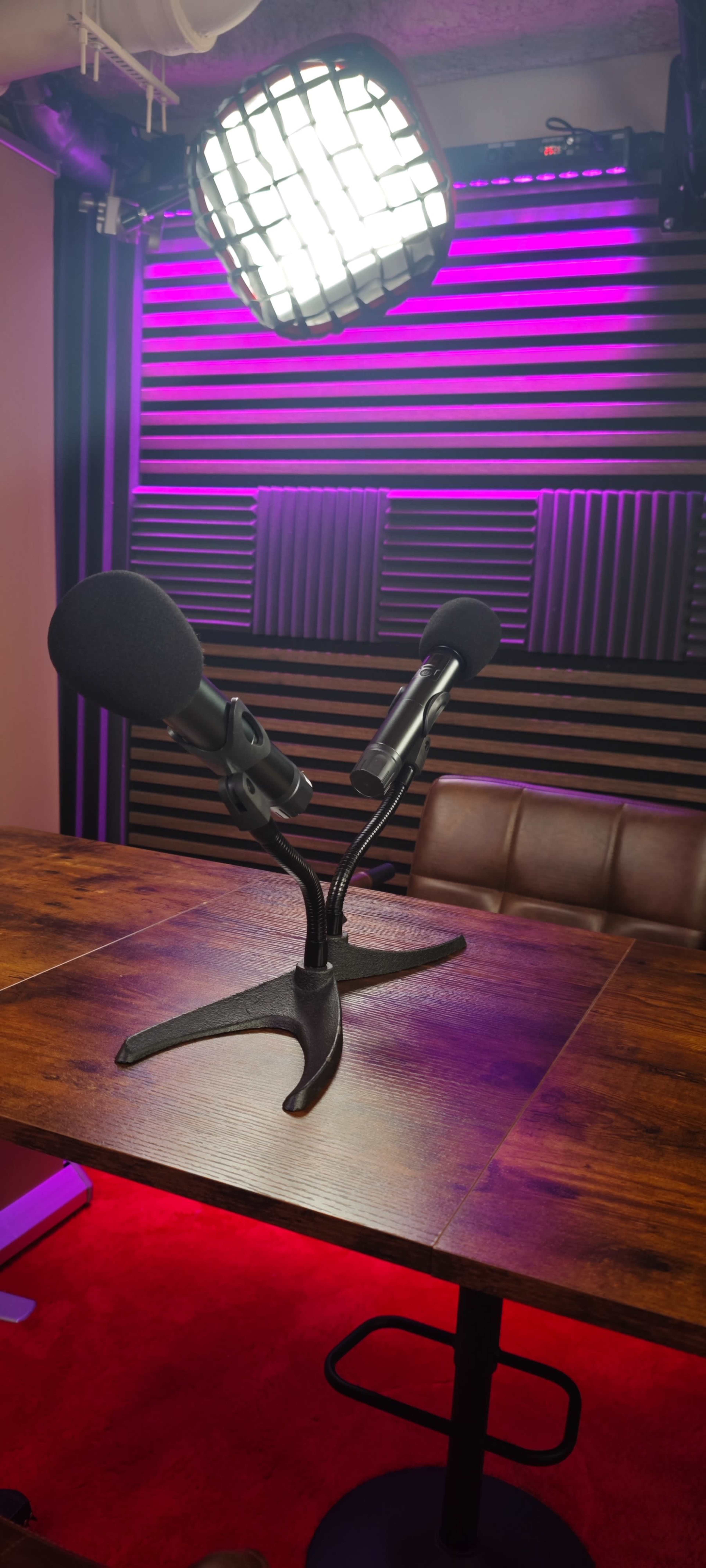 The image shows a modern podcasting setup with two microphones positioned on a wooden table in front of soundproofing panels and purple lighting.