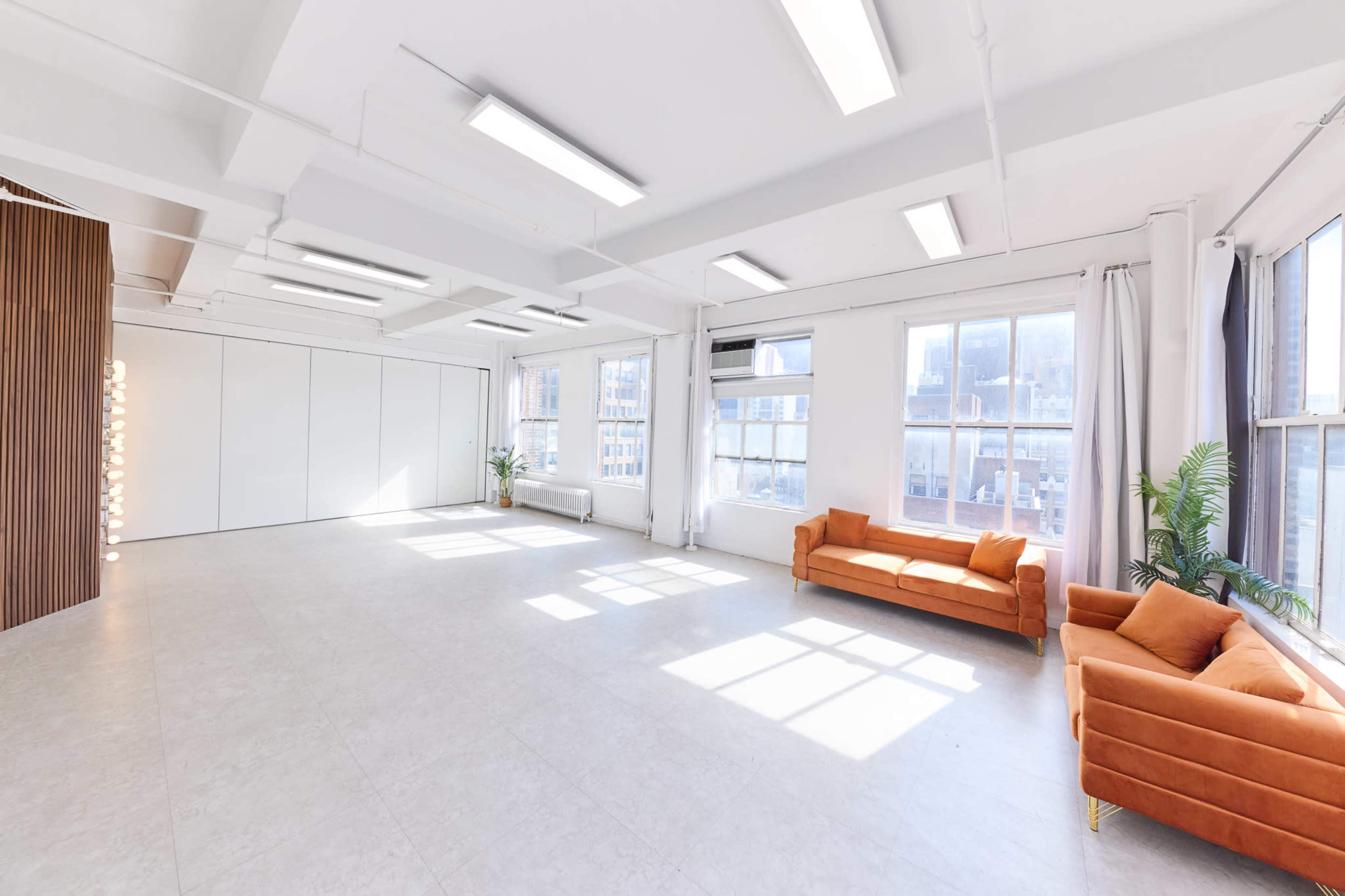 The image shows a bright, spacious room with large windows, light-colored flooring, and a pair of orange sofas.