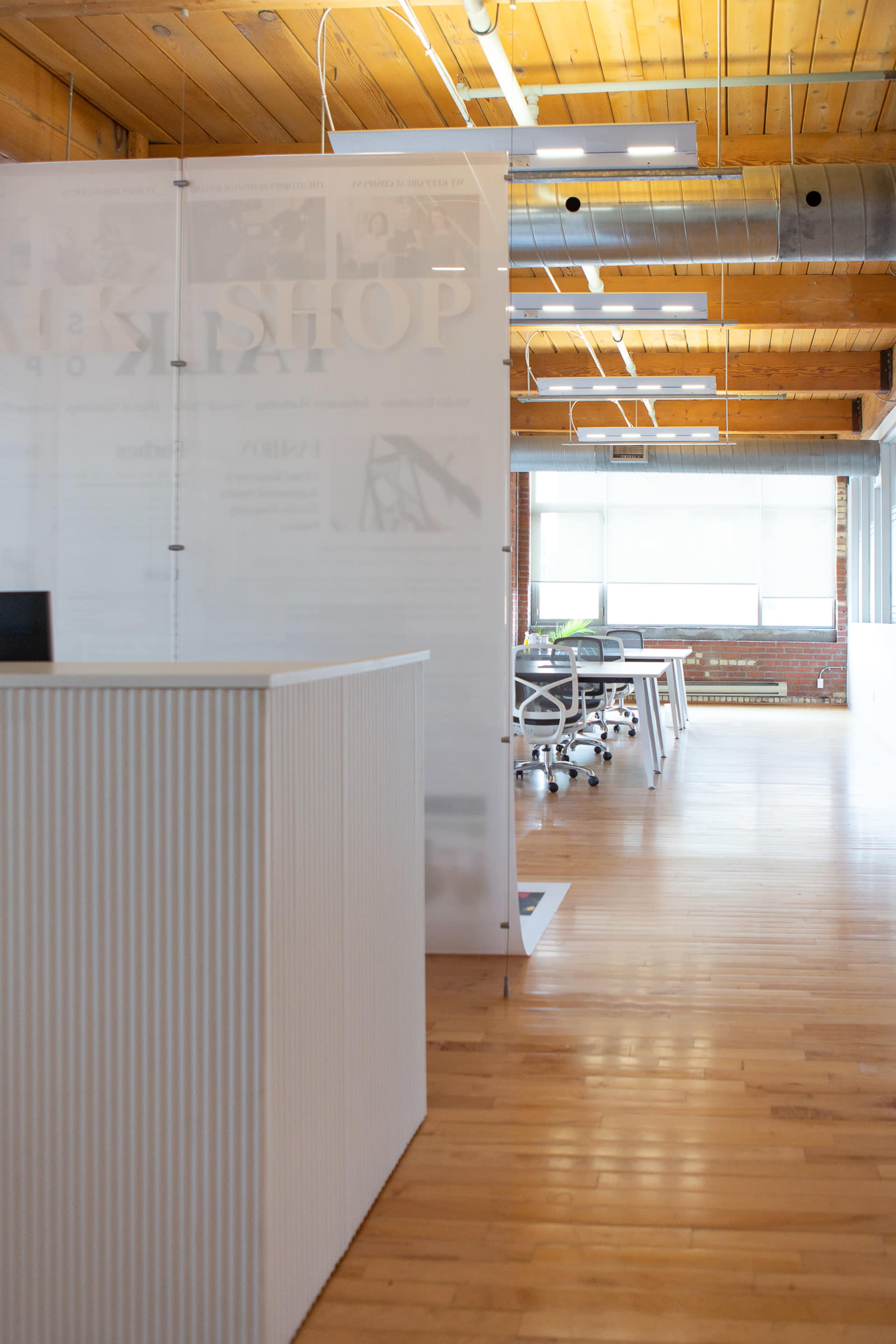 A modern office space with wooden floors, large windows, and a partition separating the reception area from the workstations.