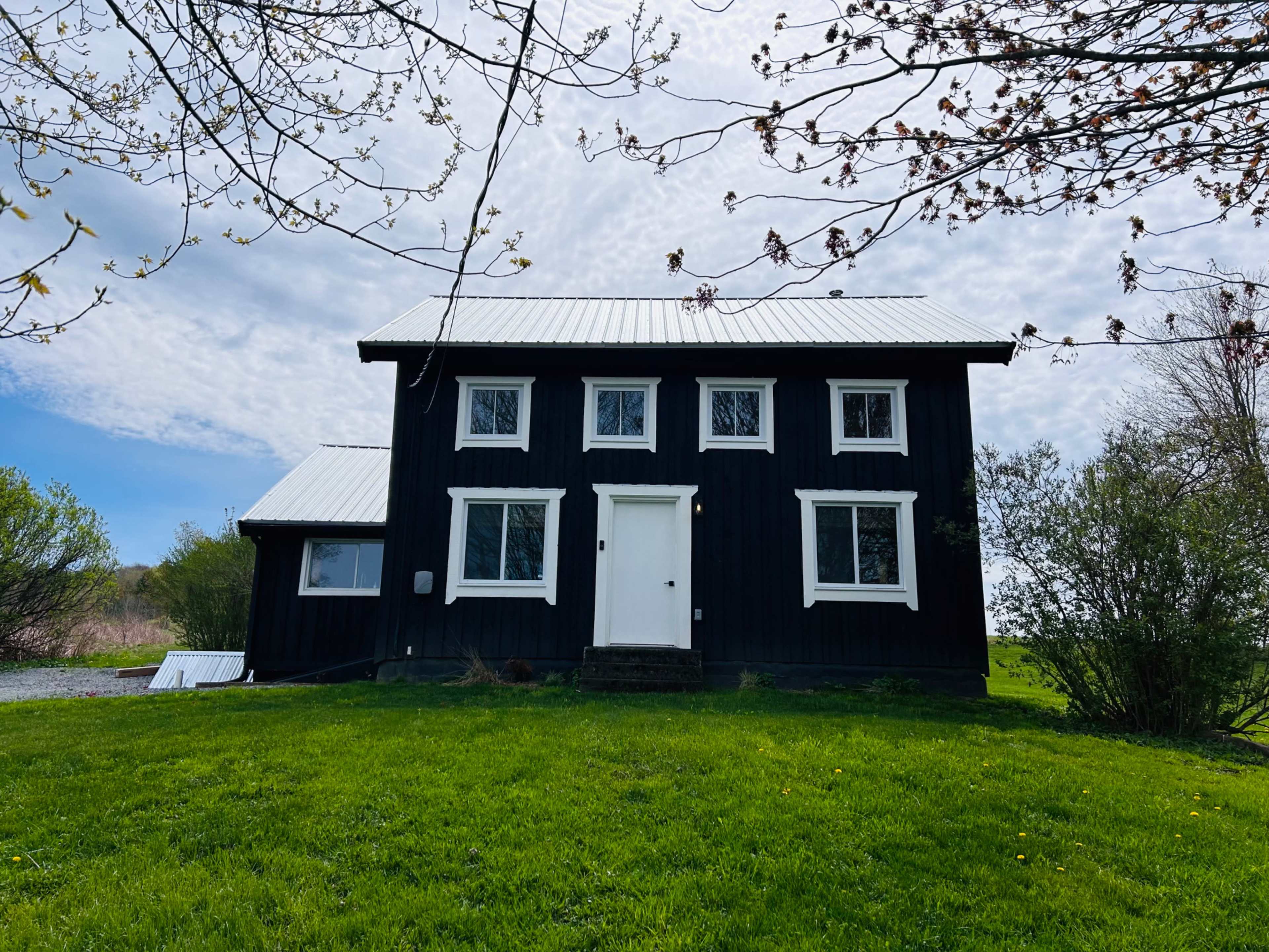A black two-story house with a white roof and several windows sits on a green lawn under a cloudy sky.