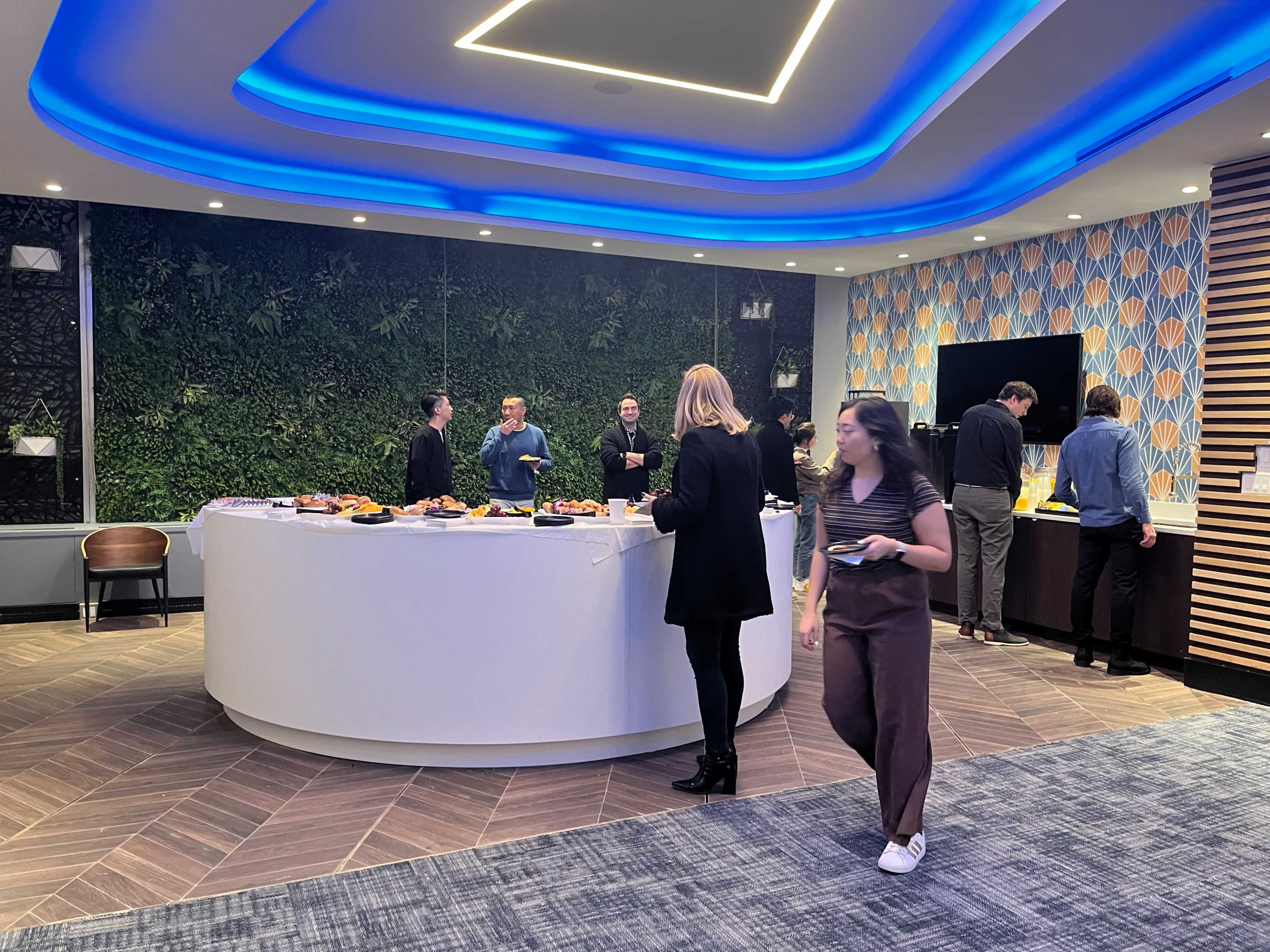 A group of people gather around a curved buffet table filled with food in a modern indoor setting adorned with greenery and decorative lighting.