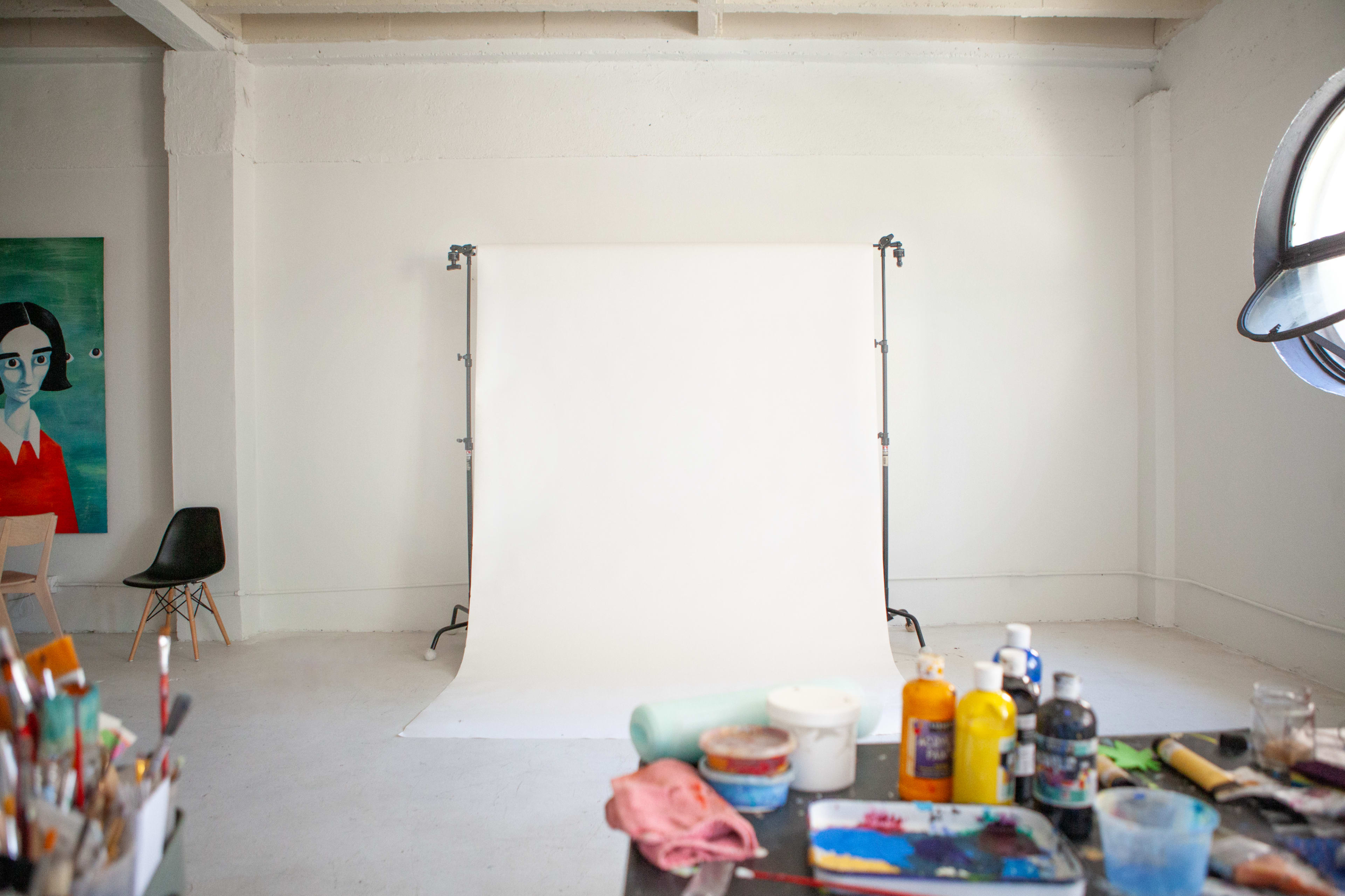 The image shows a well-lit studio space with a blank white backdrop, a chair, and art supplies scattered on a table.