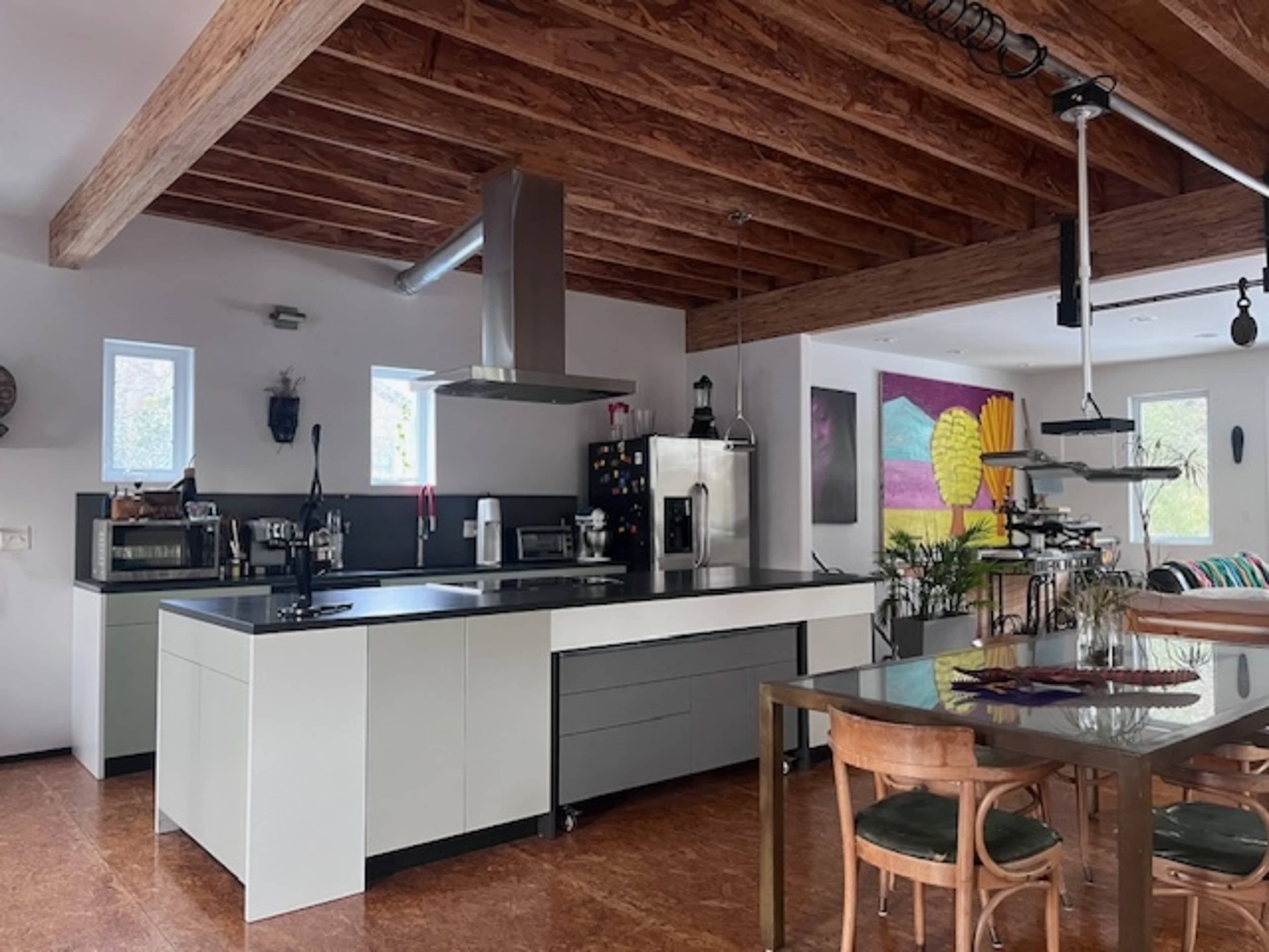 The image shows a modern kitchen with a large island, stainless steel appliances, wooden beams, and a dining area featuring a glass table and wooden chairs.