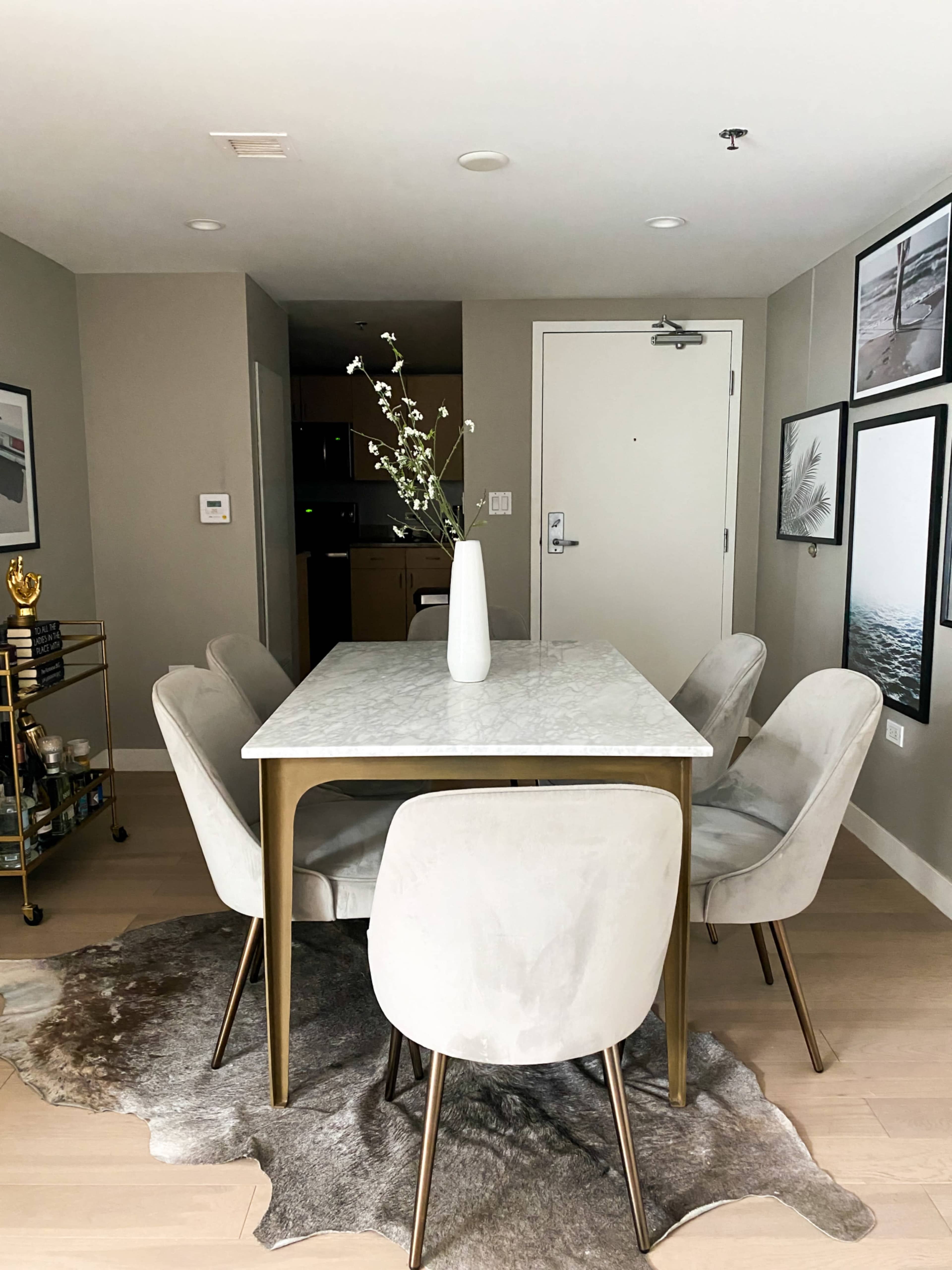 A dining area features a marble-topped table surrounded by six light-colored chairs, with a vase of flowers as the centerpiece and framed images on the wall.