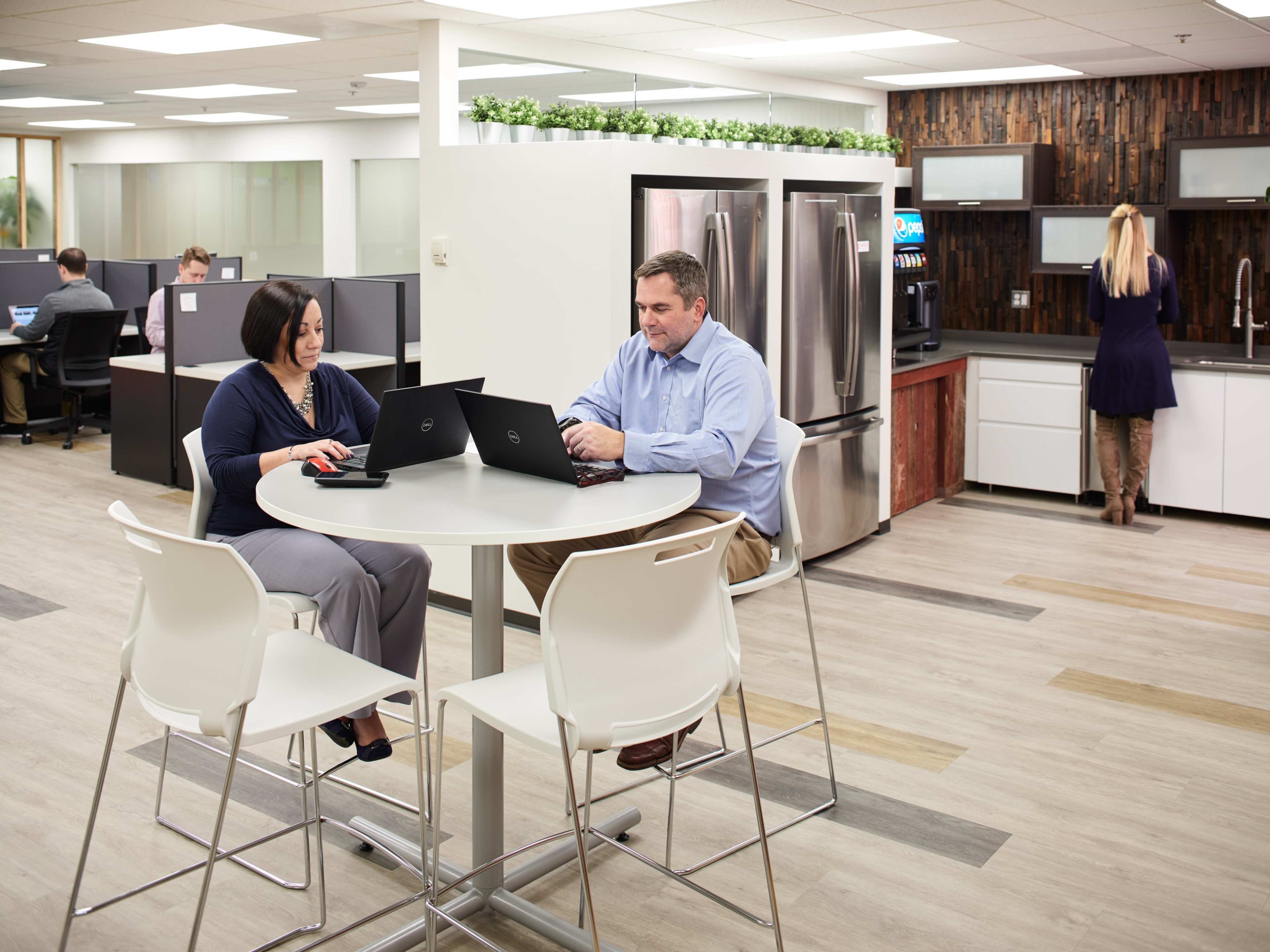 Two people work on laptops at a round table in a modern office kitchen area, while a third person stands at the sink.