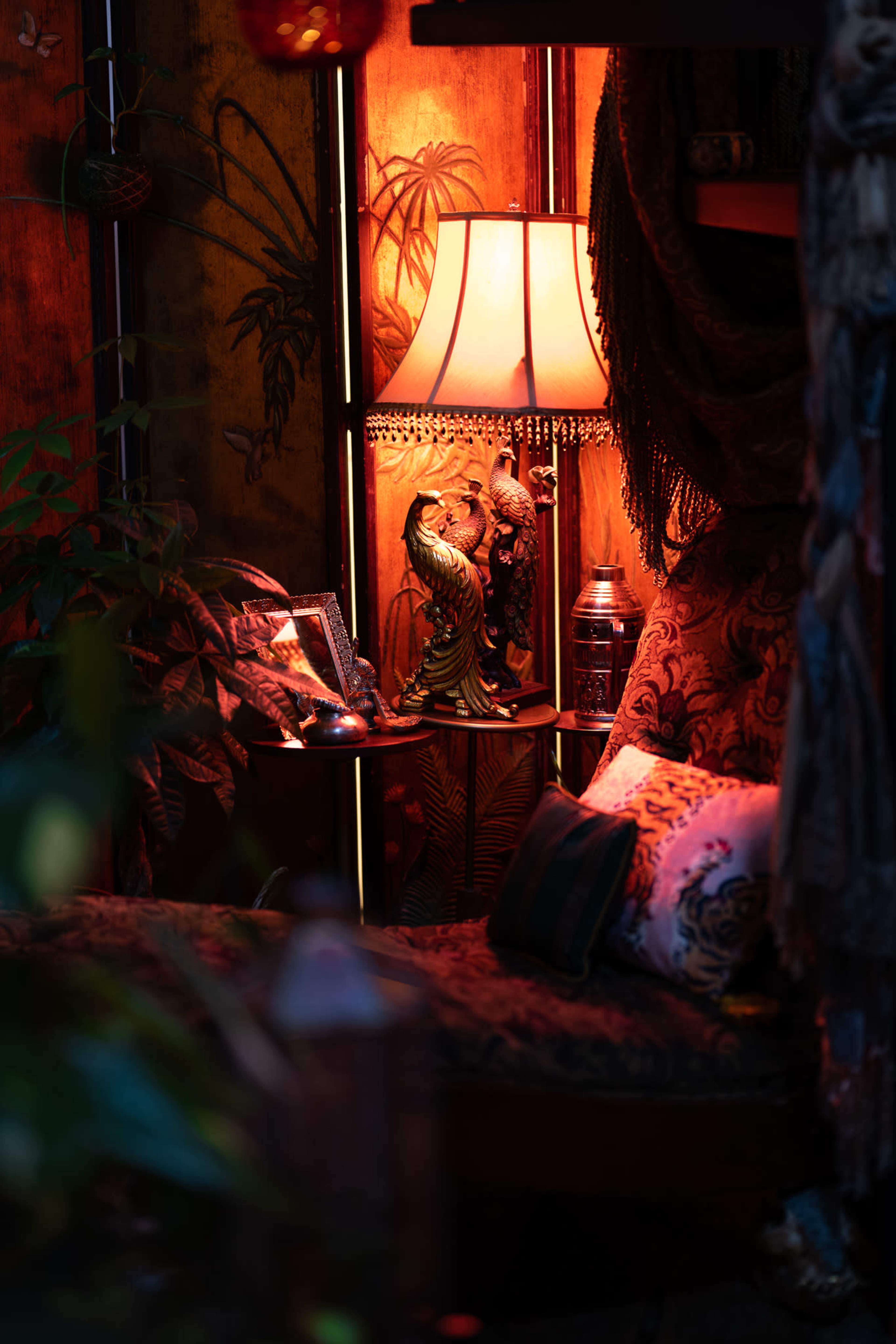 The scene features a cozy corner with a lamp illuminating a patterned couch, surrounded by plants and decorative items.