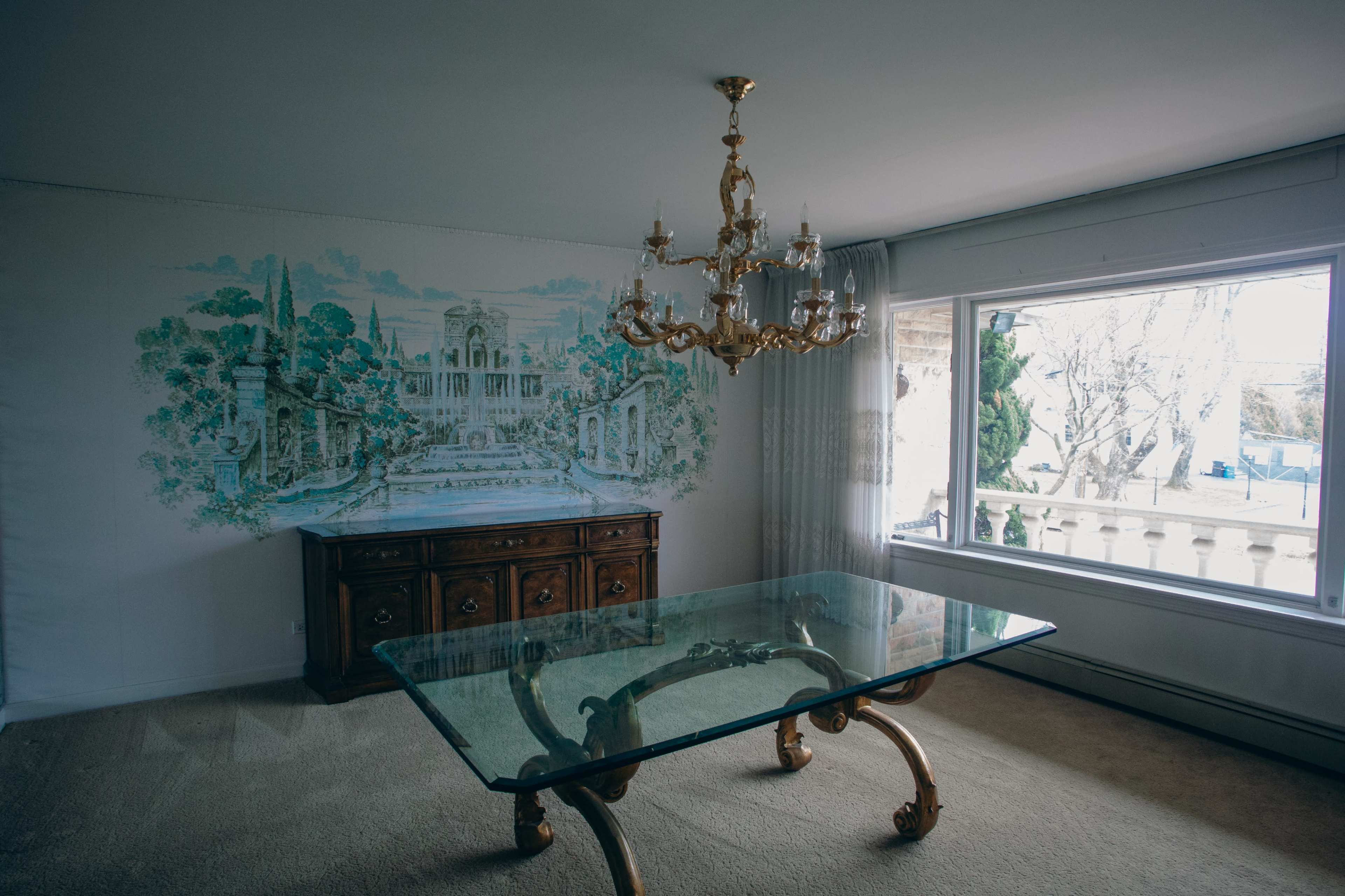 The image shows a dining room with a glass table, a wooden sideboard, a chandelier, and a mural on the wall depicting a scenic landscape.