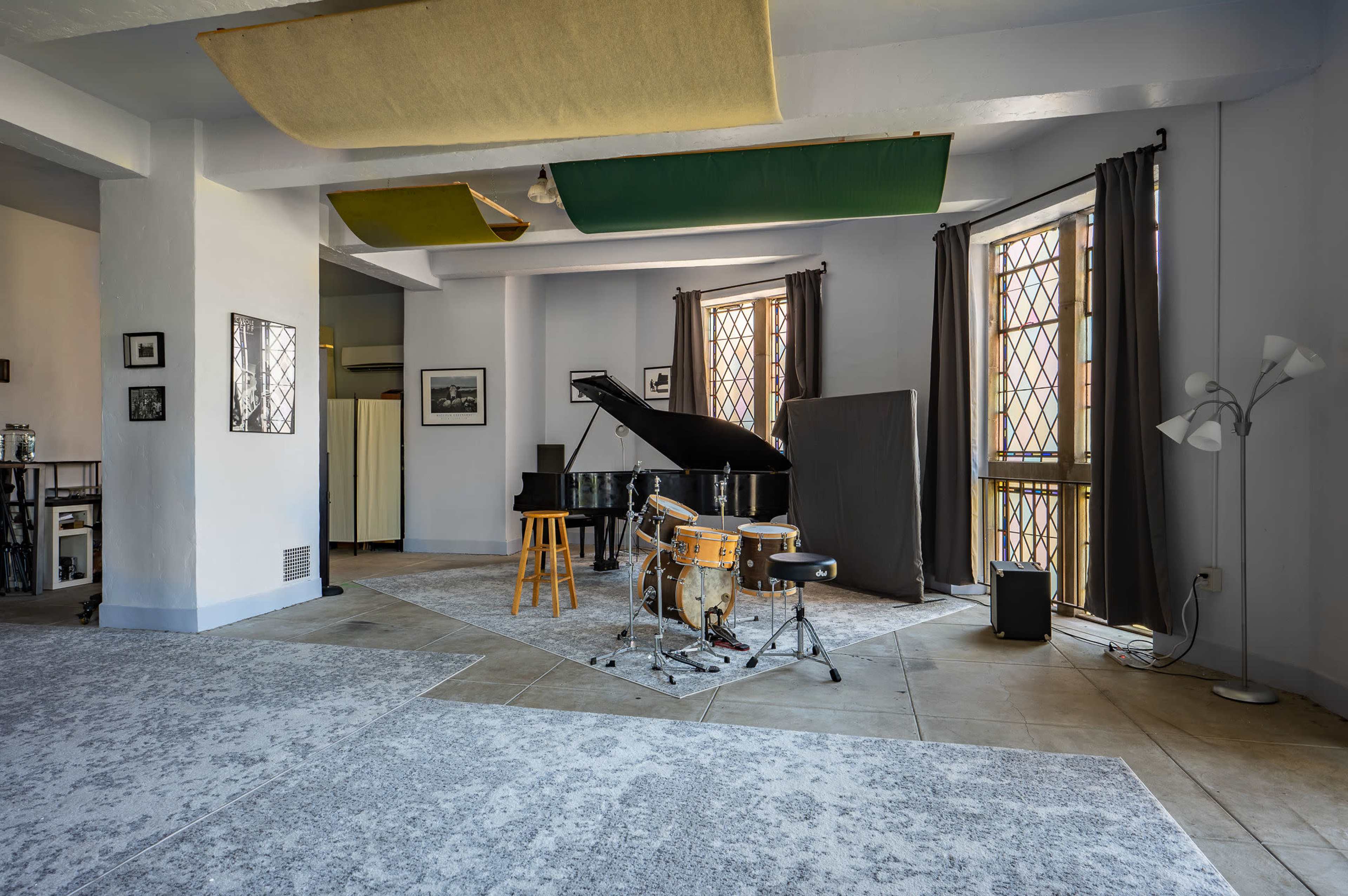 The image shows a music room featuring a grand piano, a drum set, and large windows with stained glass, all set on a carpeted floor.