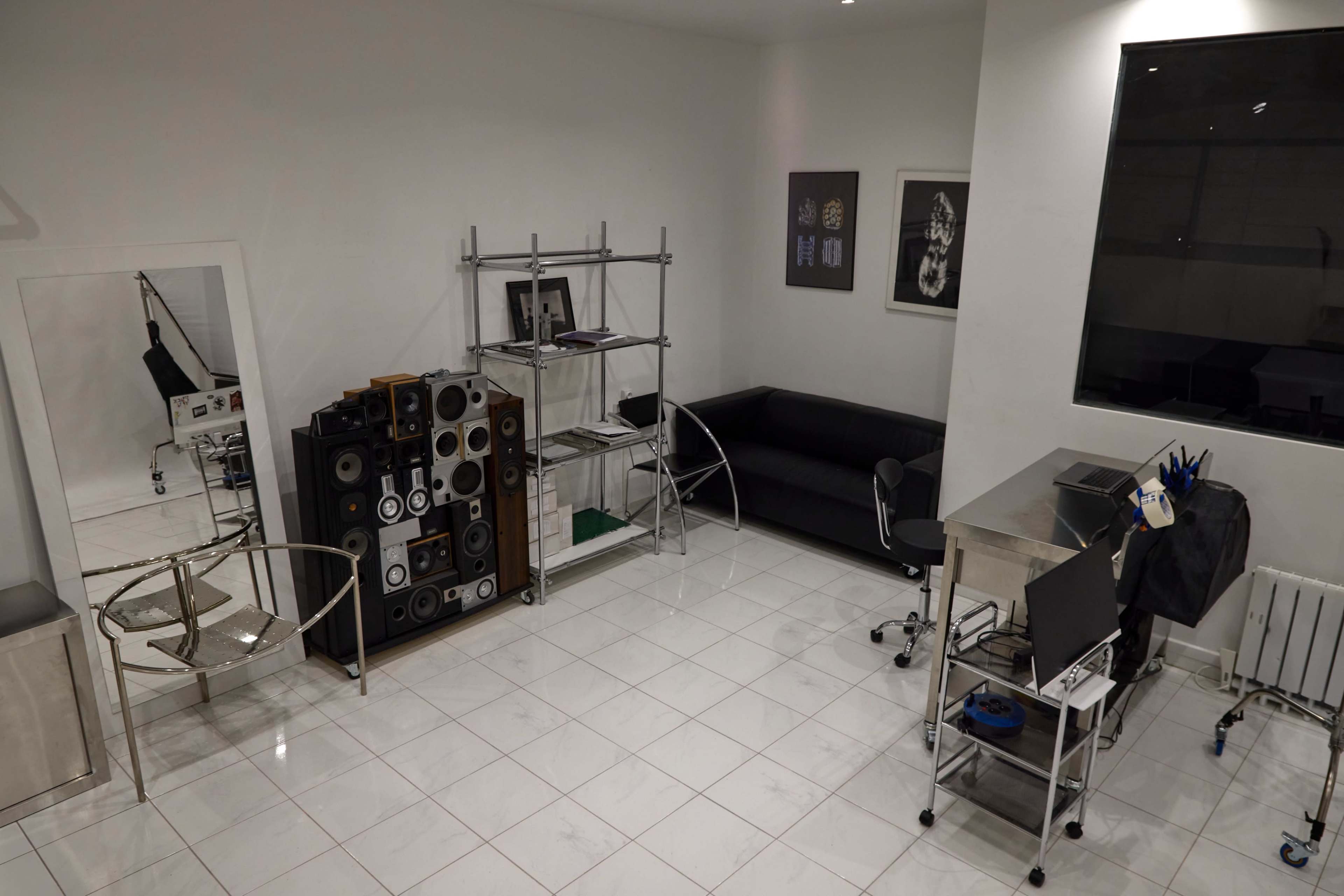 The image shows a modern, minimalist room featuring a black couch, a metal shelf with speakers, a glass table, and a workstation with a computer.