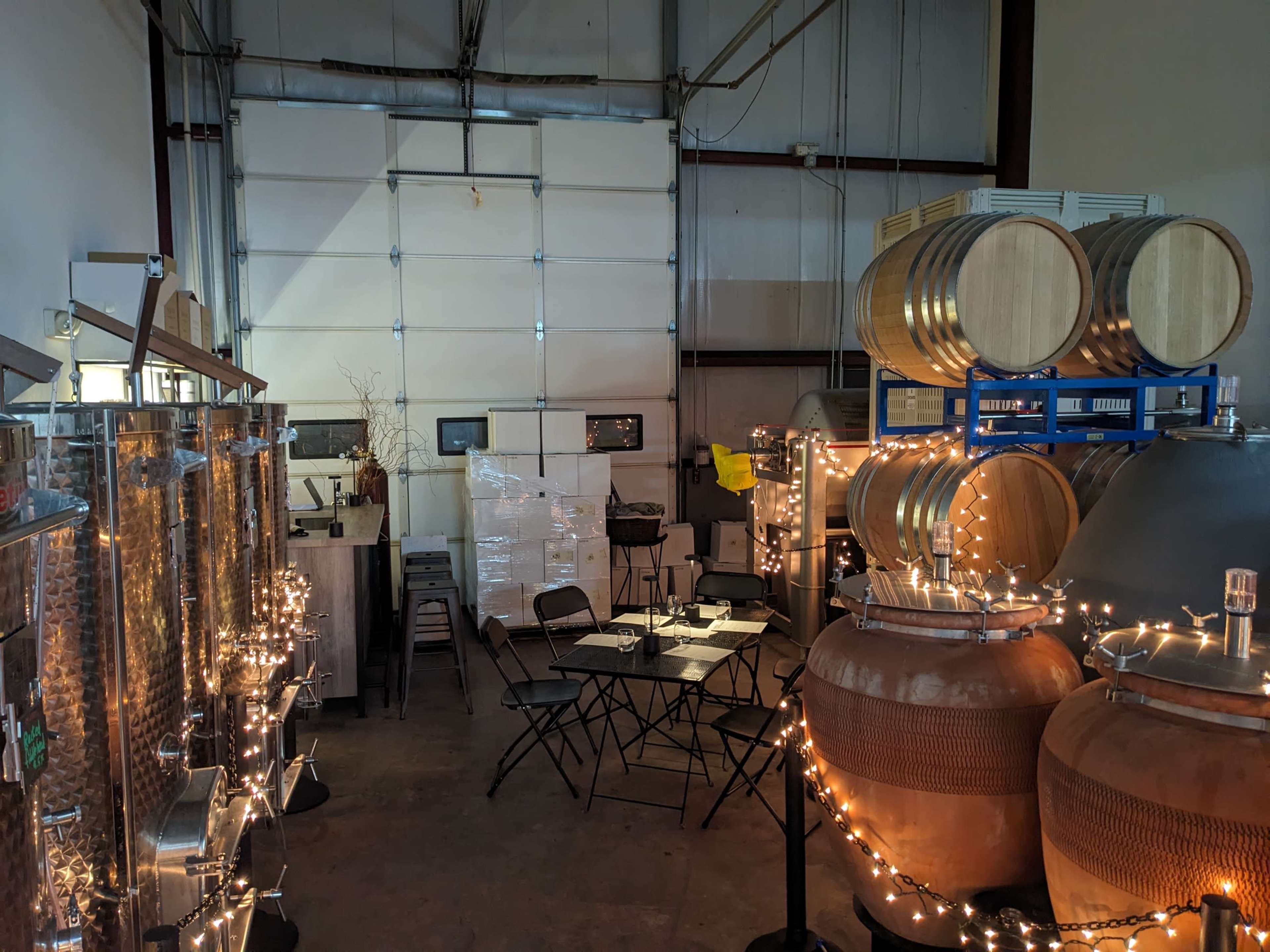 A spacious winery interior with large fermentation tanks and barrels, a few tables set up, and string lights illuminating the area.