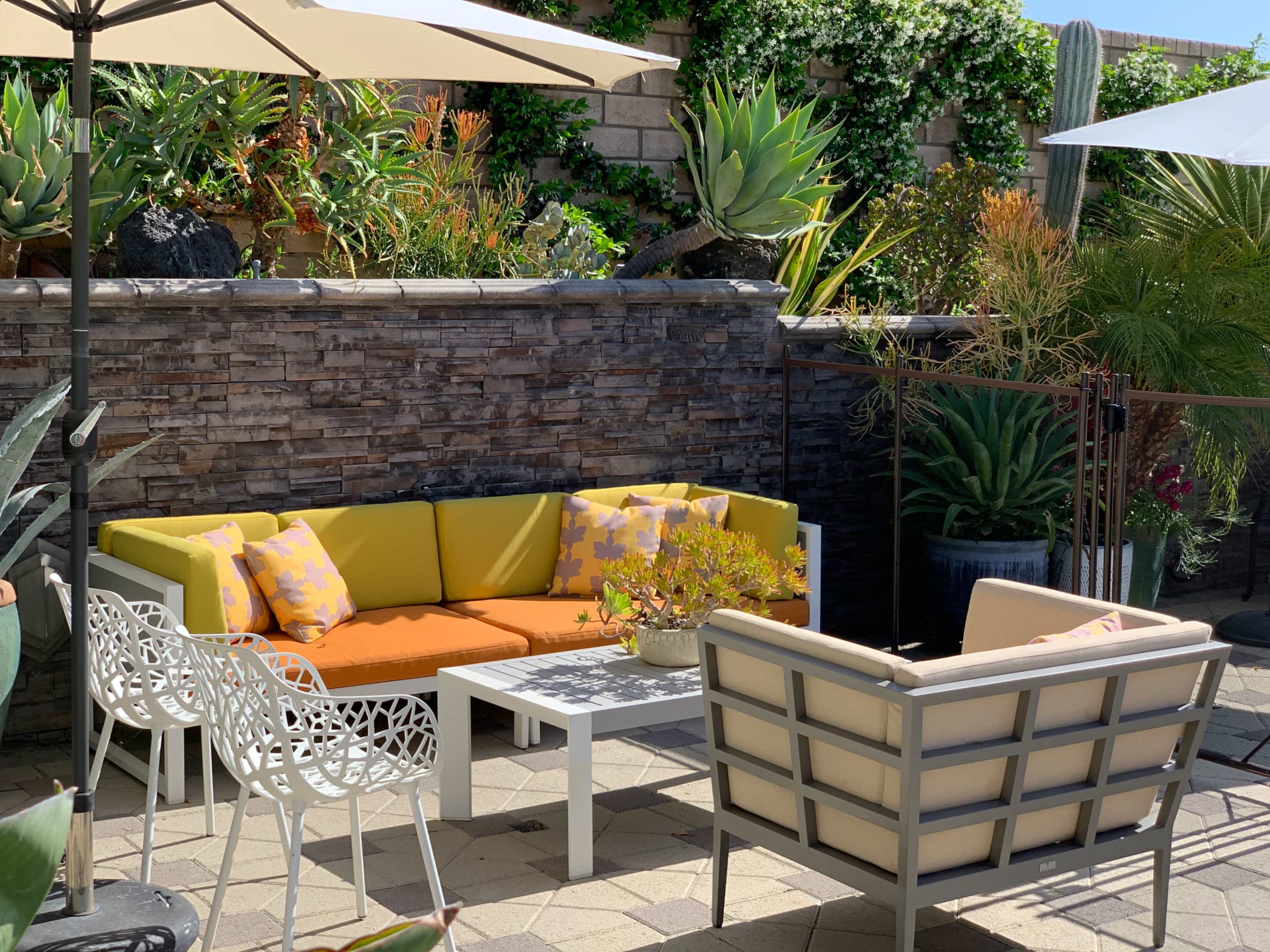 A colorfully furnished outdoor seating area features a modern couch, a coffee table, and decorative plants against a stone wall.