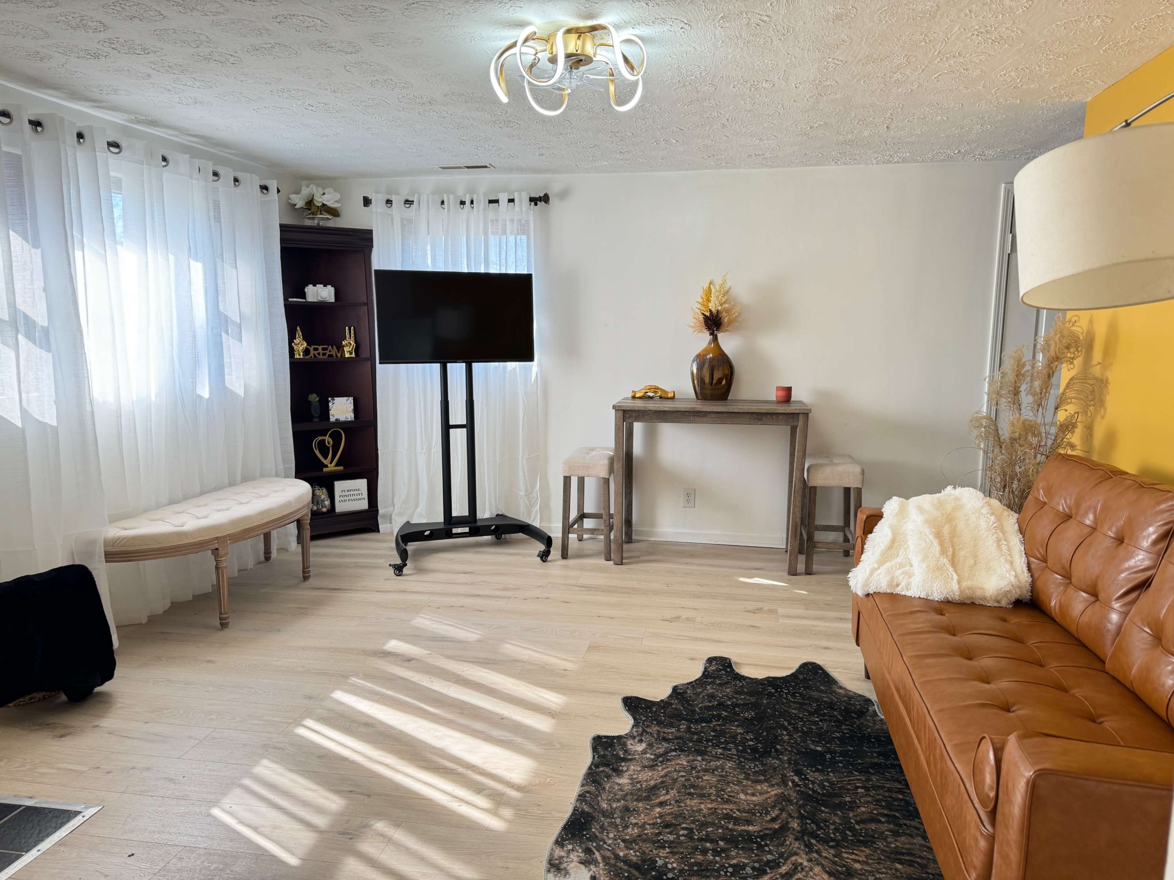 A living room with a brown leather sofa, a small table, a television on a stand, and large white curtains letting in natural light.
