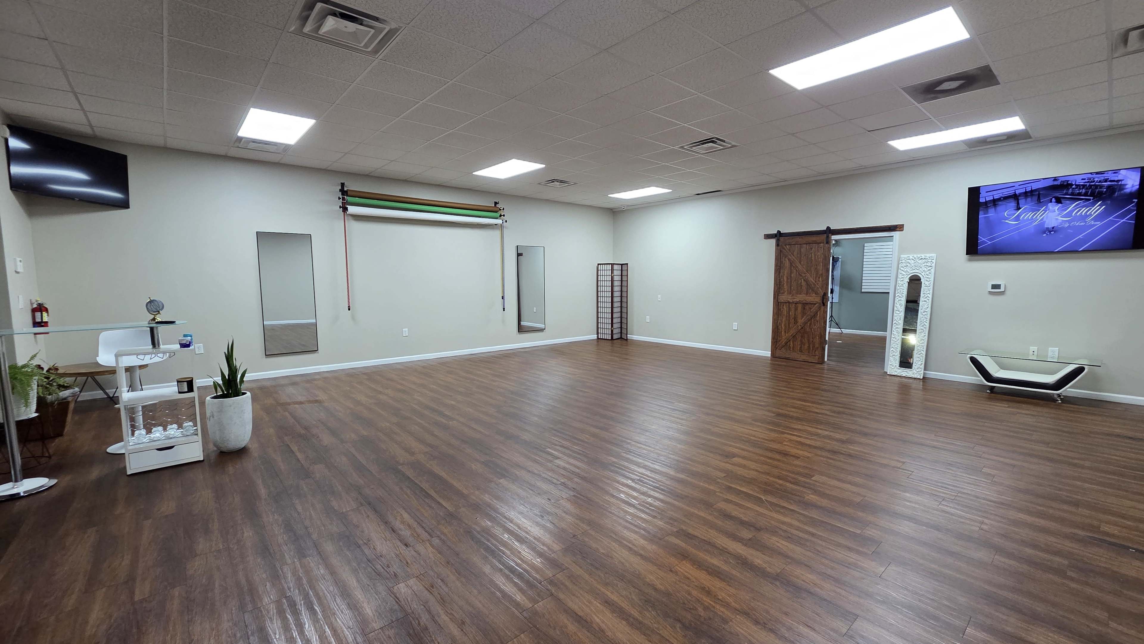 The image shows a spacious, empty room with wooden flooring, bright overhead lights, and mirrored walls, featuring a sliding barn door and a large screen on one wall.