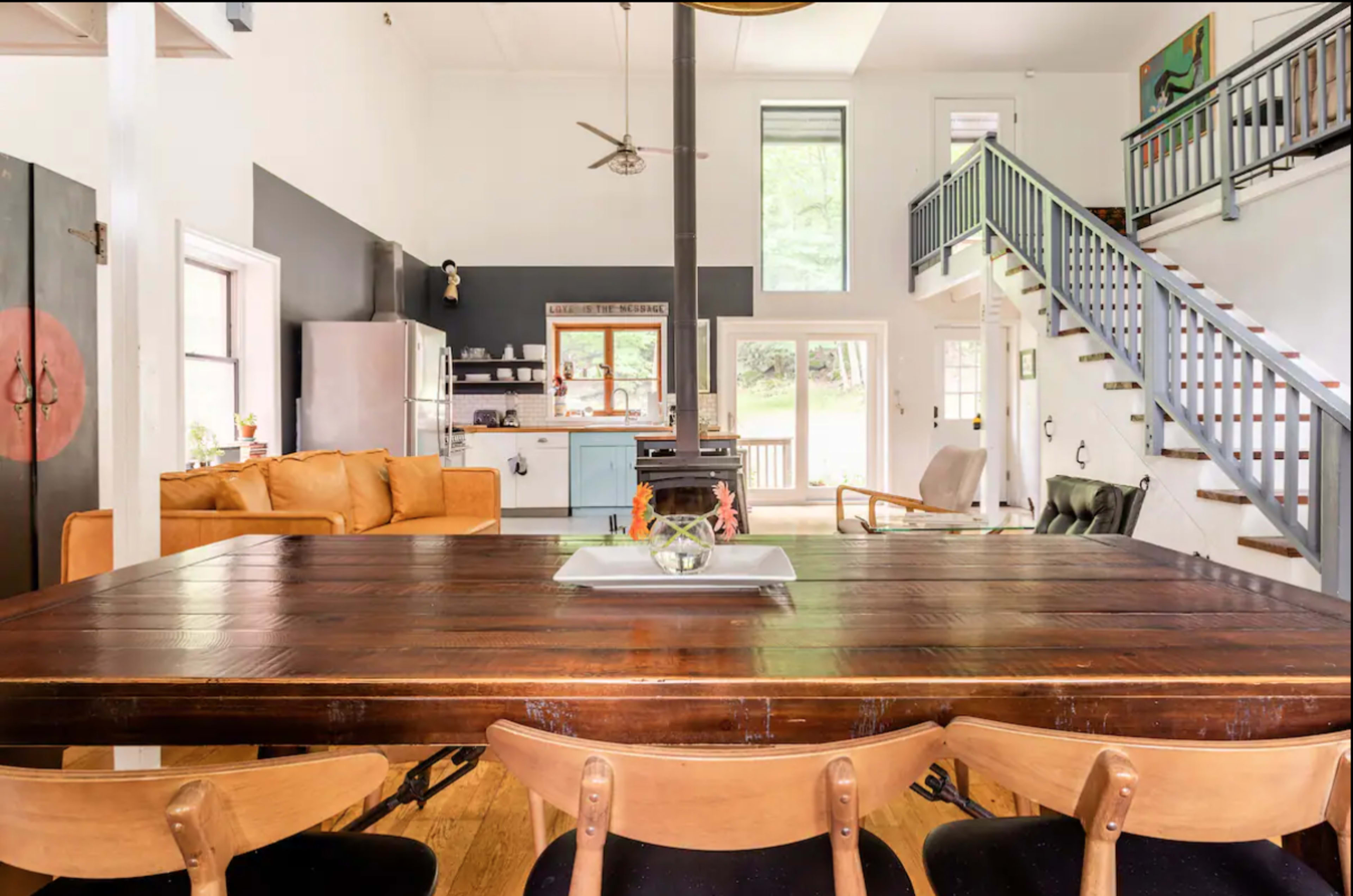 The image shows a modern open-concept living space with a wooden dining table in the foreground, a kitchen in the background, and stairs leading to an upper level.