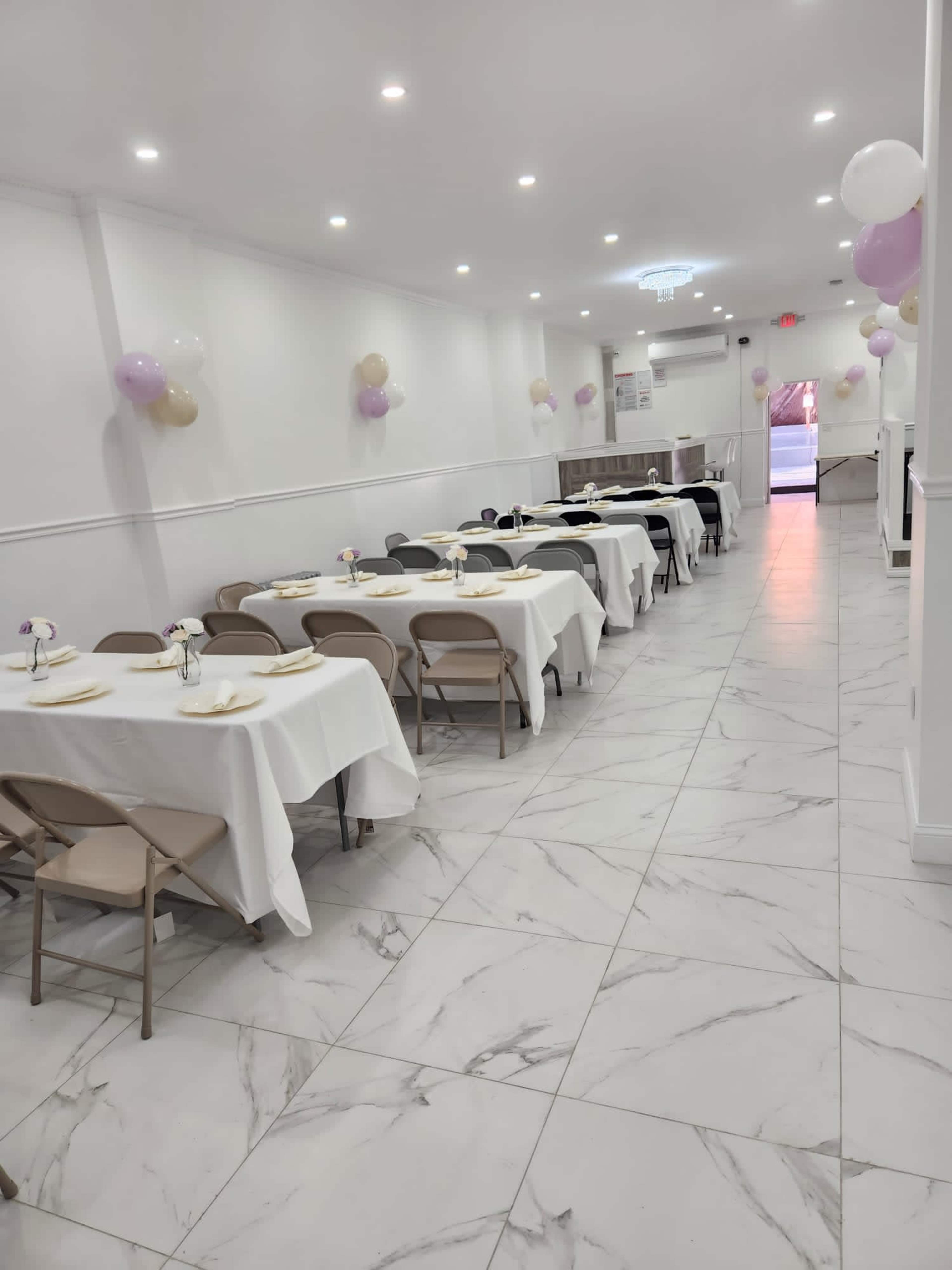 A long, narrow room set up for an event, featuring neatly arranged tables covered in white tablecloths and decorated with small floral centerpieces.