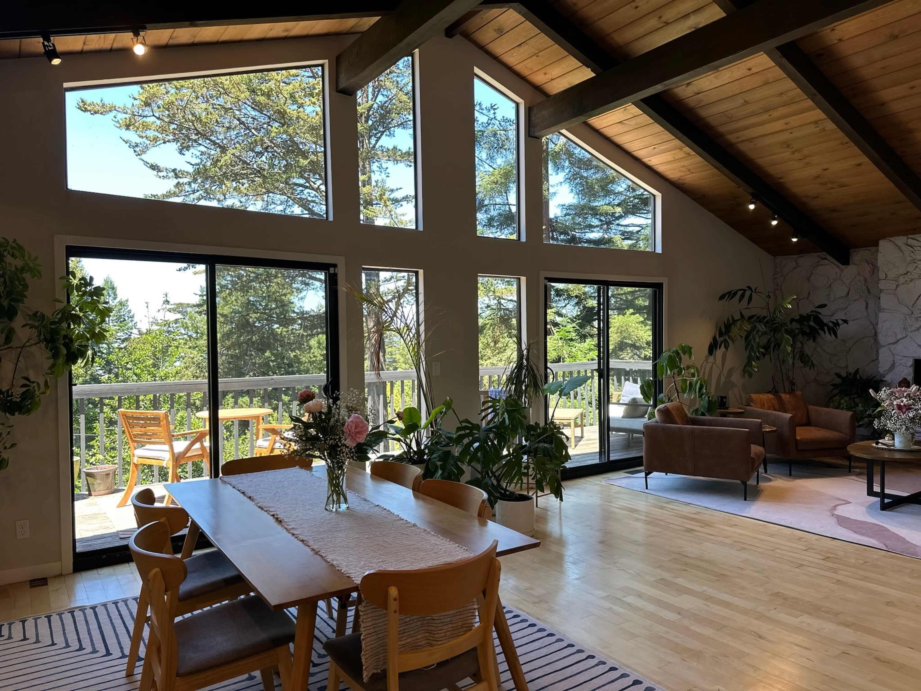 A spacious living area features large windows overlooking a deck surrounded by trees, with a dining table and plants placed throughout the space.