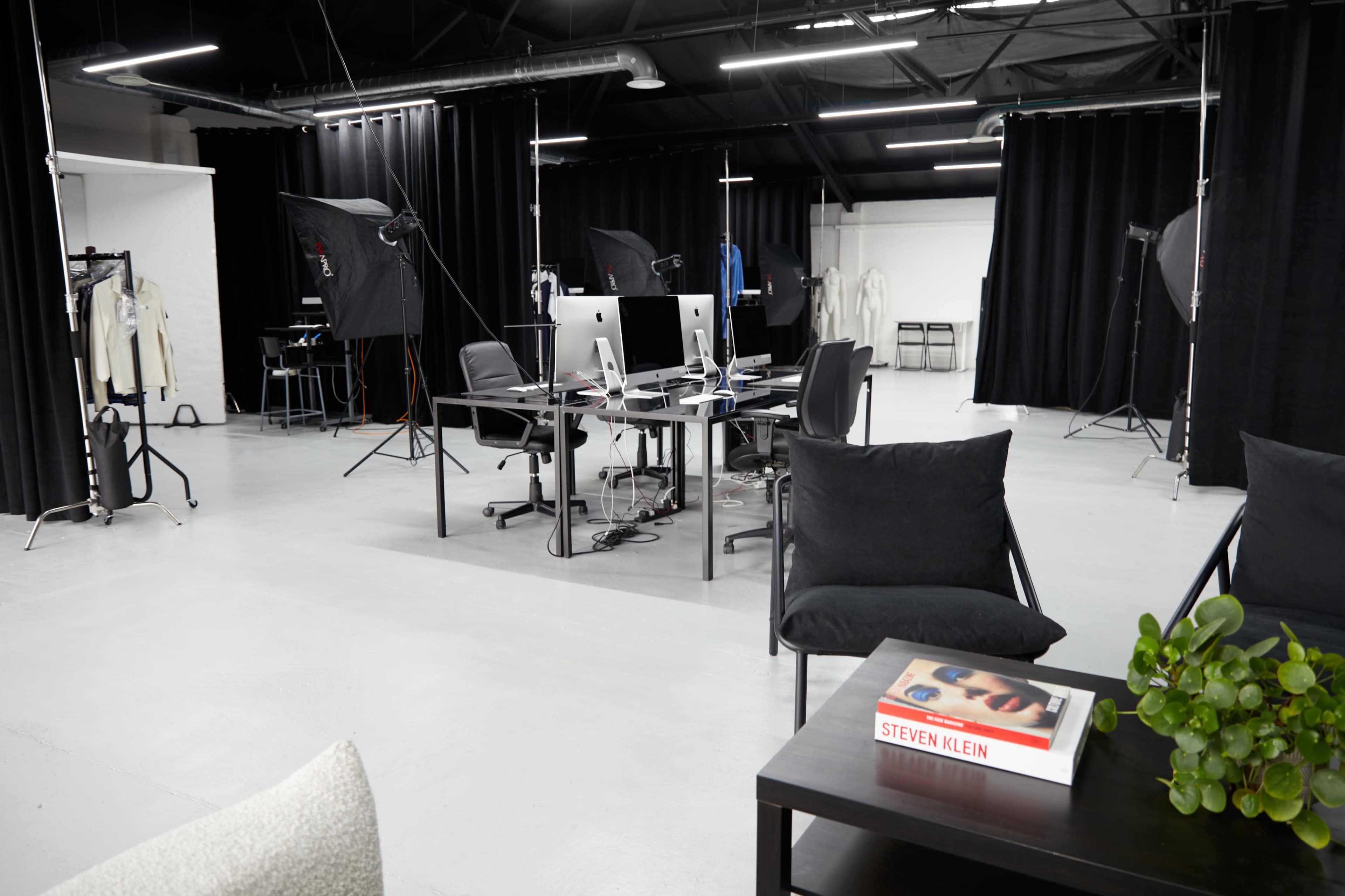 The image shows a photography studio with several computers on desks, chairs, and various lighting setups, all surrounded by black curtains.