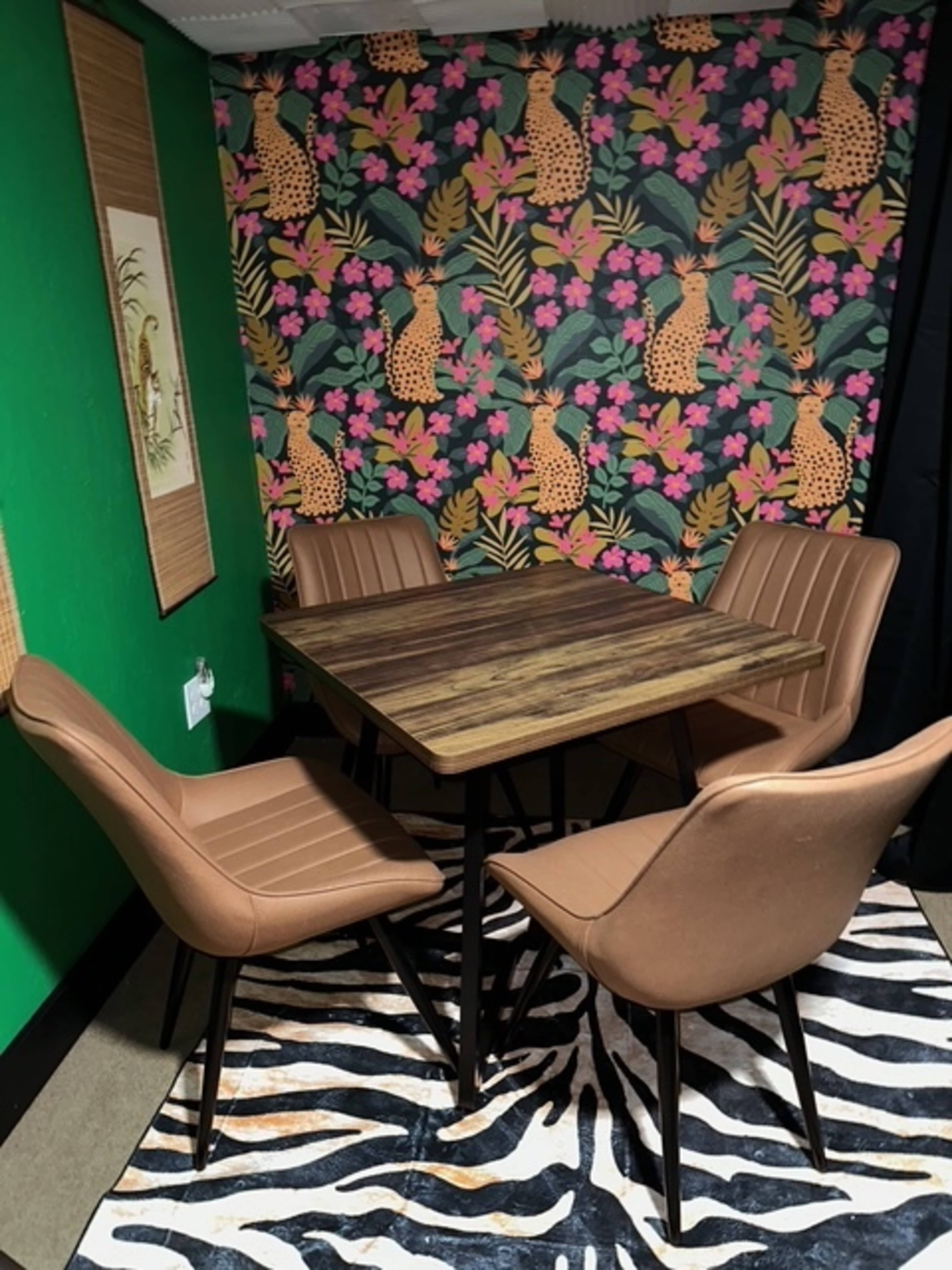 The image shows a small dining area featuring a square wooden table surrounded by four brown upholstered chairs, set against a colorful floral and animal-patterned wallpaper.
