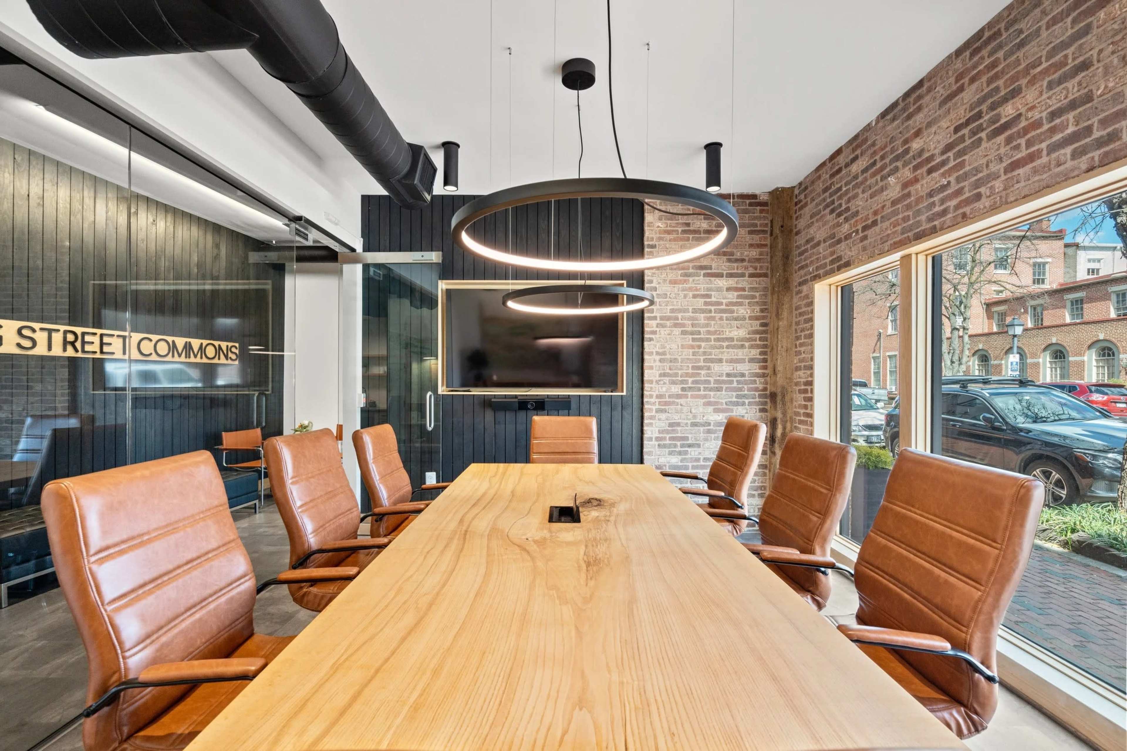 A modern conference room features a large wooden table surrounded by brown leather chairs, with a circular light fixture overhead and a large screen on the wall.