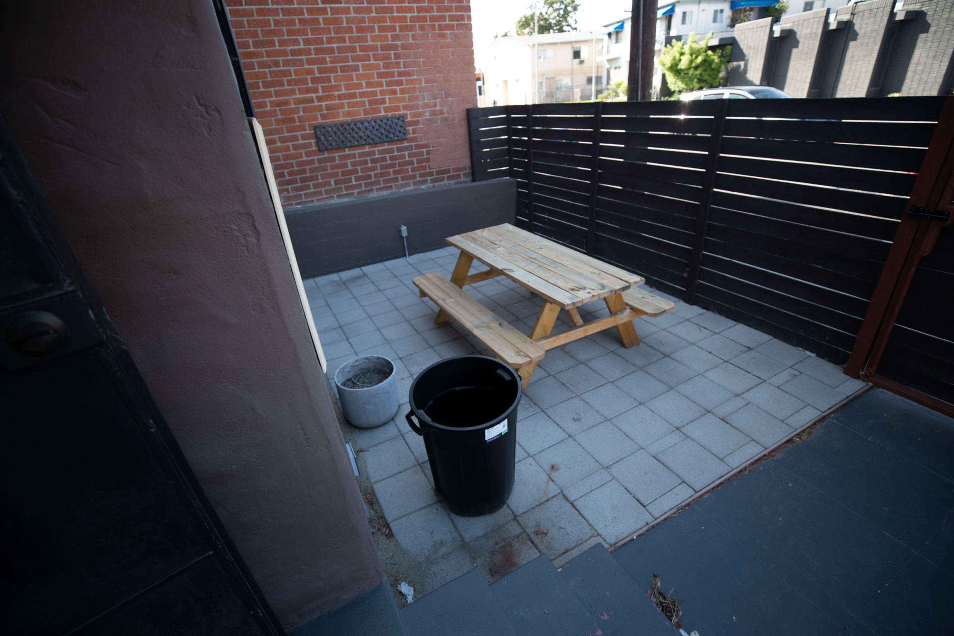 A small outdoor space featuring a wooden picnic table, a black trash can, and a potted plant on a tiled surface, enclosed by a wooden fence and a brick wall.