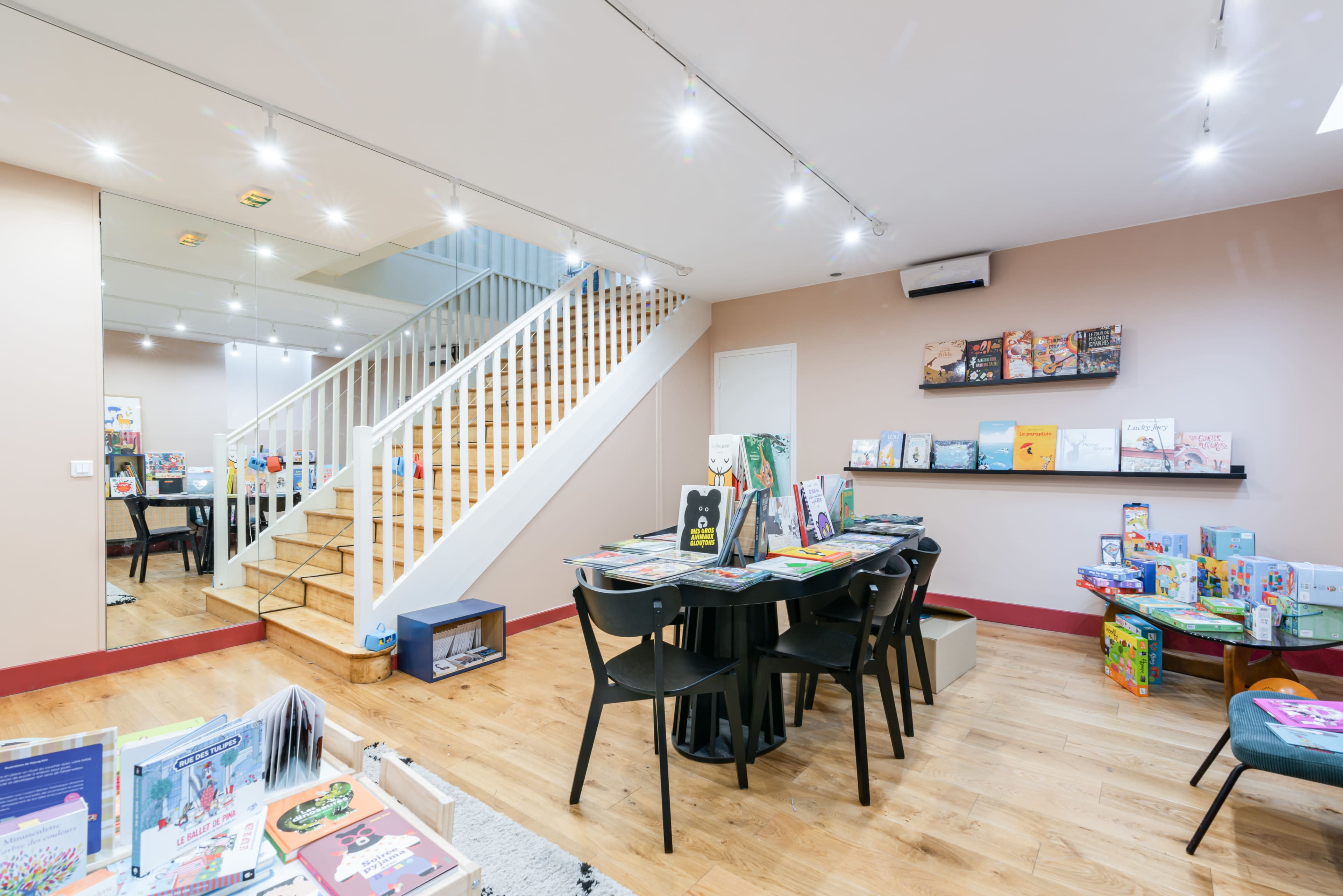 A brightly lit children's bookstore features a central table with books and games, surrounded by wooden shelves and a staircase in the background.
