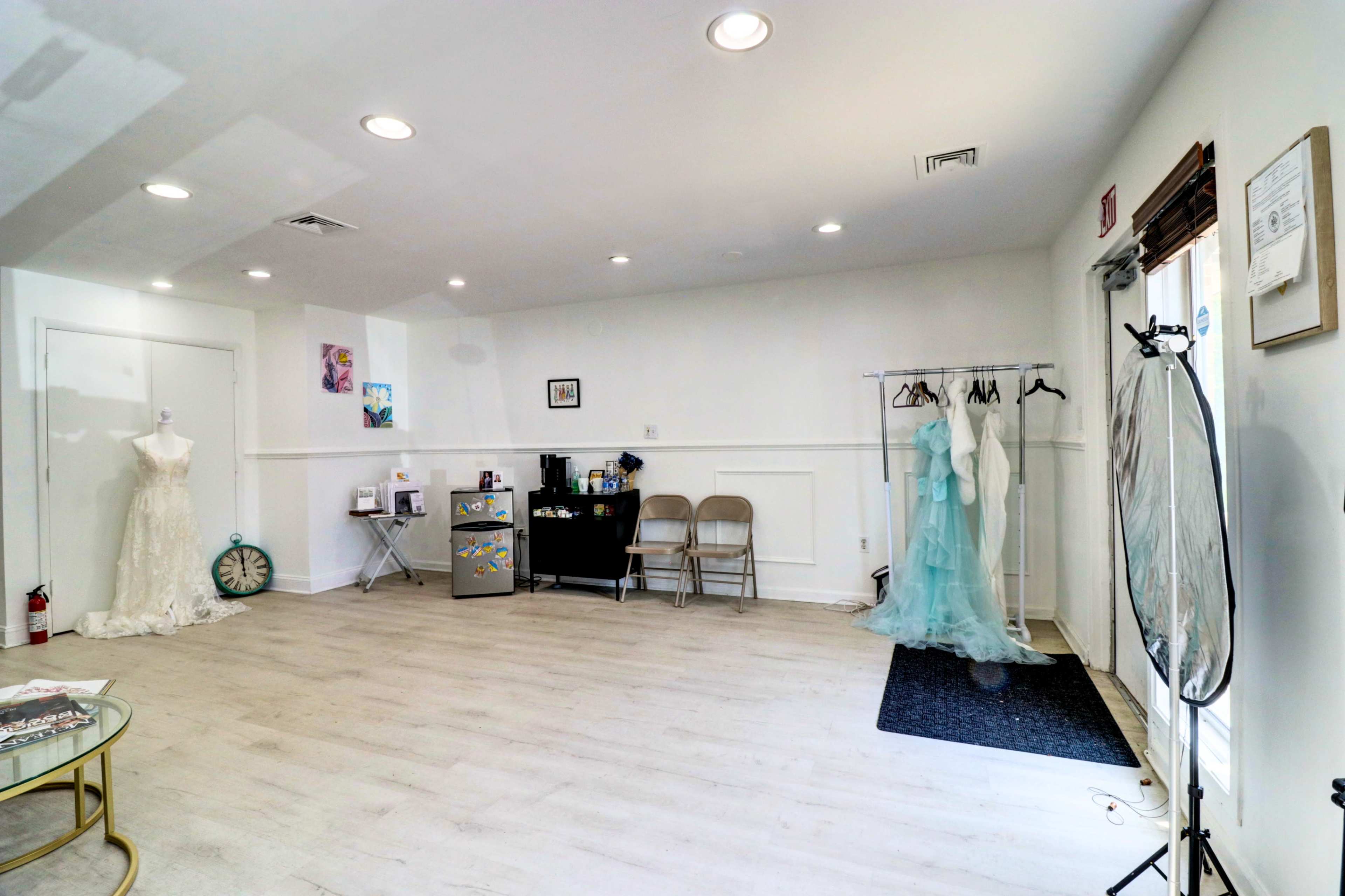 The image shows a bright, empty studio space with a mannequin wearing a wedding dress, a clothing rack with several dresses, a small table, and a few chairs.