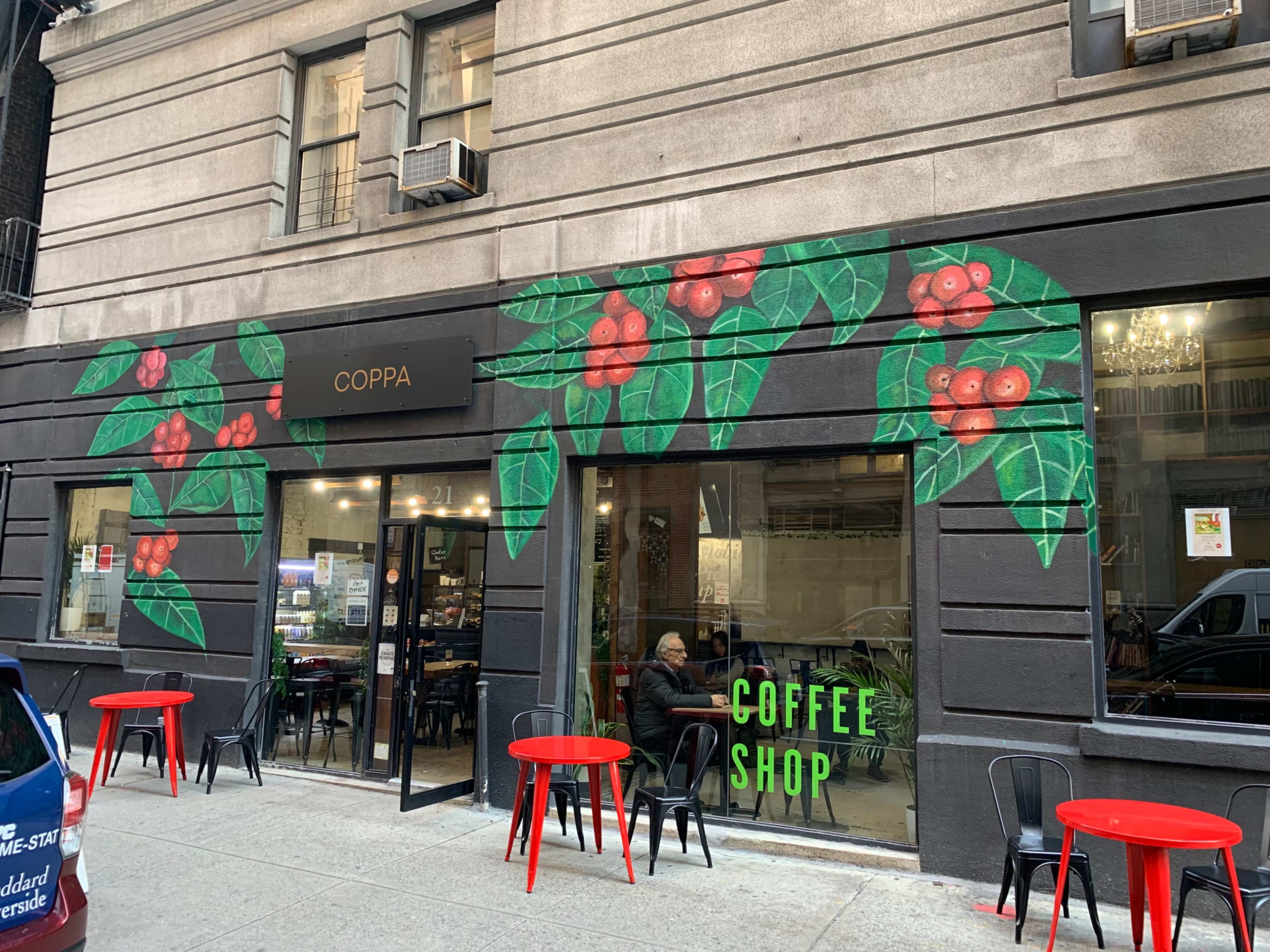 A coffee shop named "Coppa" with a mural of coffee plants on the exterior wall and red tables outside.