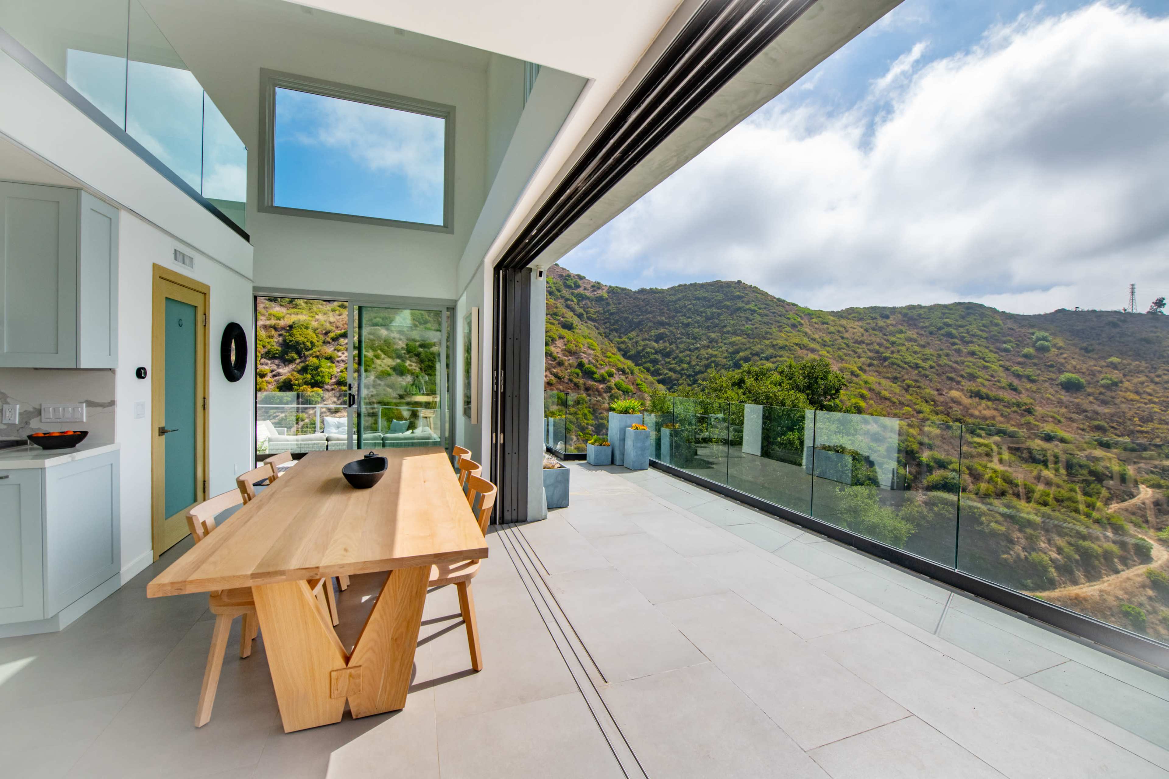 A modern dining area features a wooden table and chairs, with large glass doors that open to a view of green hills and cloudy skies.
