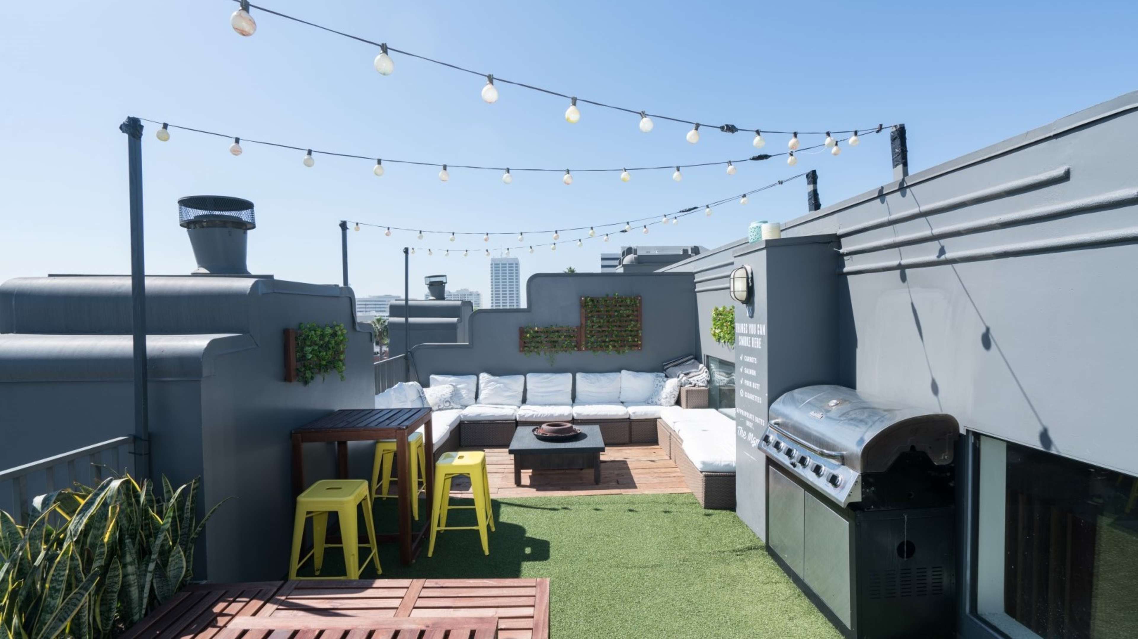 A rooftop patio with seating, a barbecue grill, string lights, and green plants against a clear blue sky.