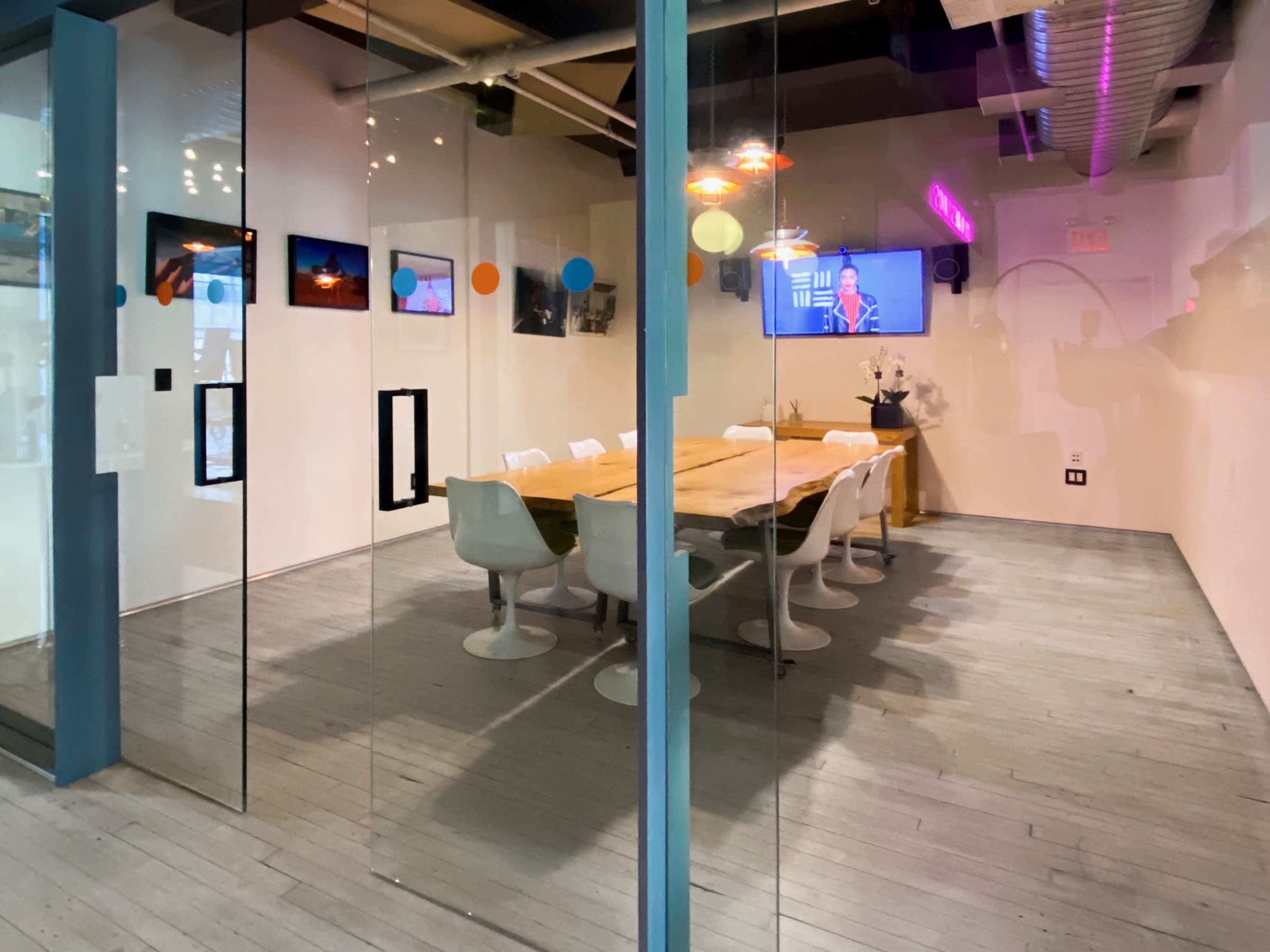 A conference room with a large wooden table and white chairs is visible through glass walls, featuring artworks on the walls and a television displaying a colorful screen.