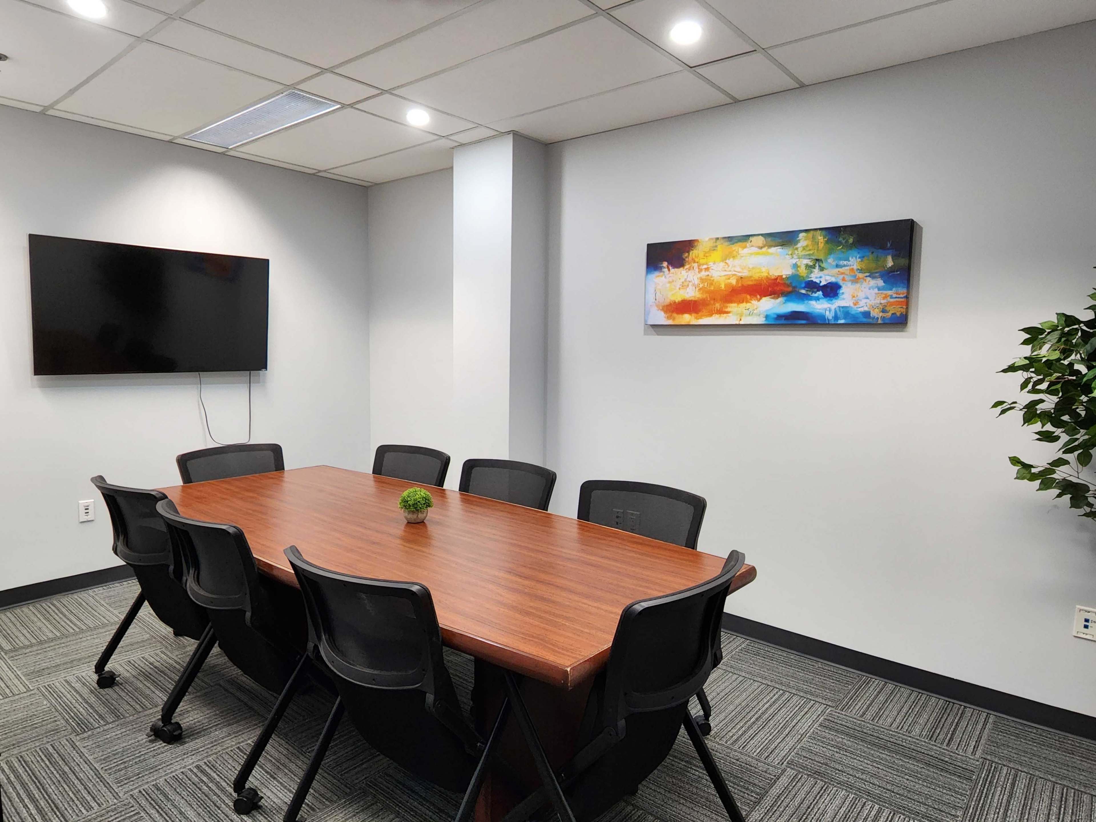 A conference room features a rectangular wooden table surrounded by black chairs, a television mounted on the wall, and a colorful artwork hanging above.