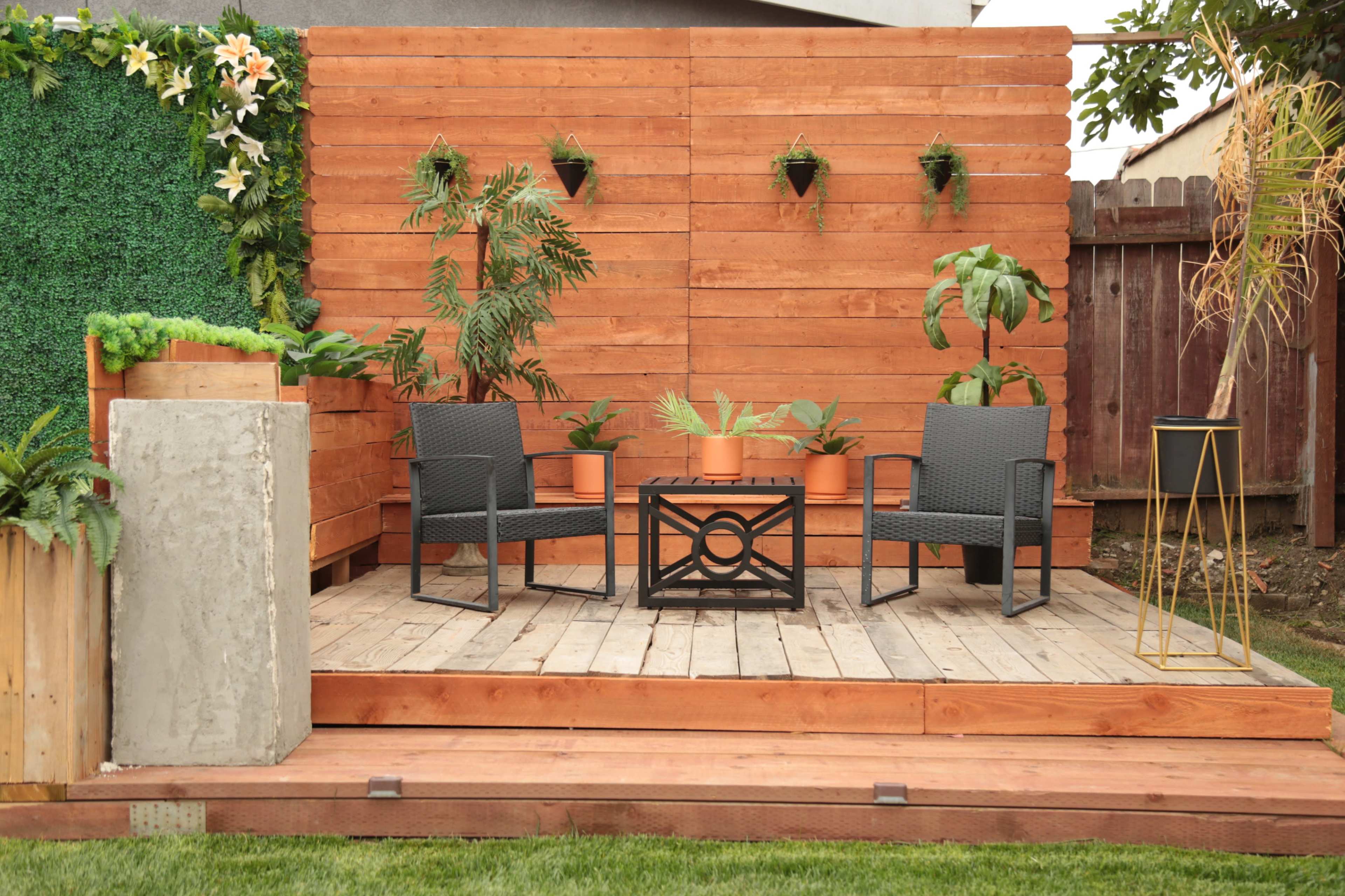 A wooden deck features two chairs and a table surrounded by potted plants against a backdrop of a wooden wall and greenery.