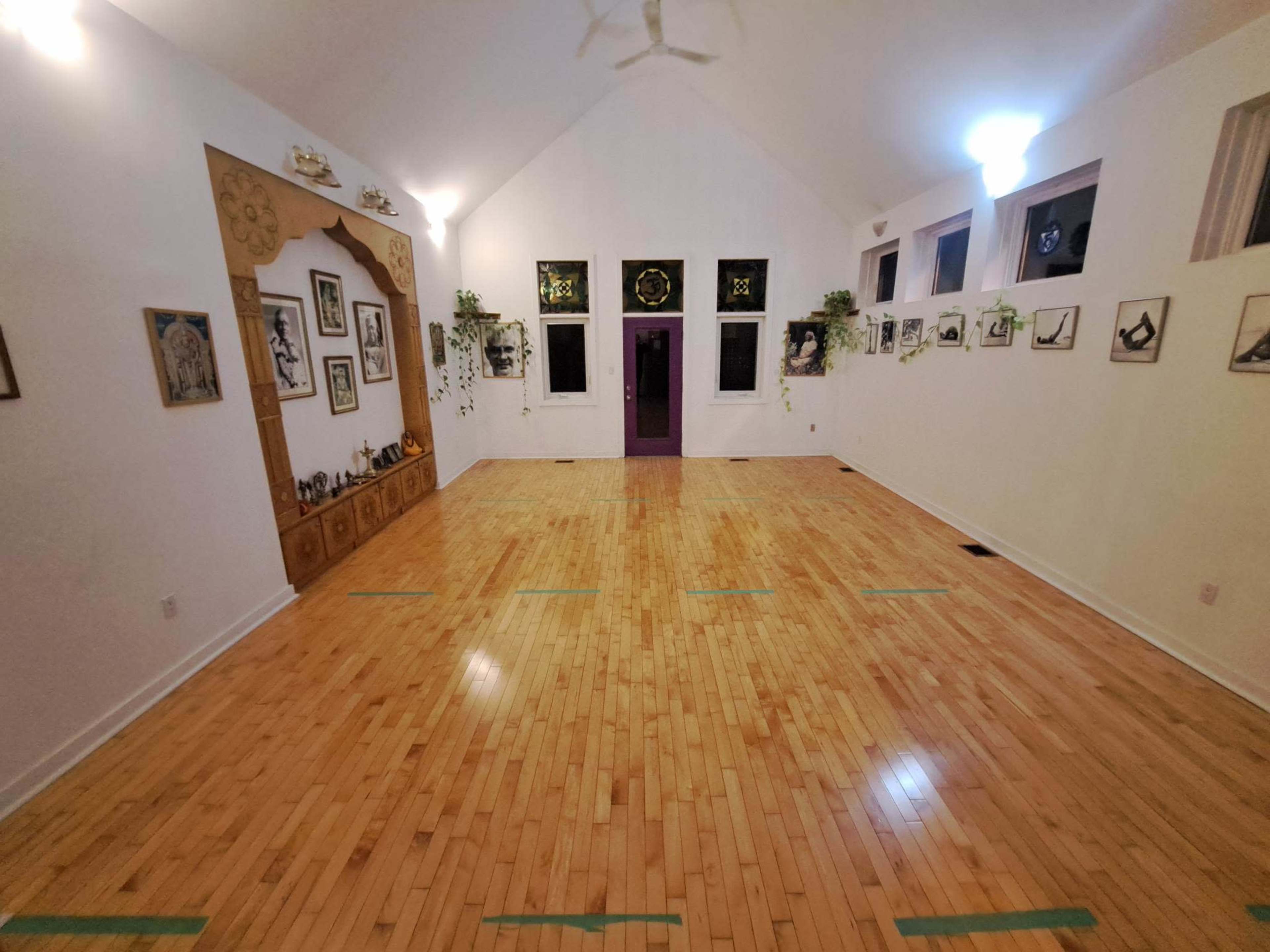 A spacious, empty interior room with wooden flooring, framed artwork on the walls, and large windows letting in light.