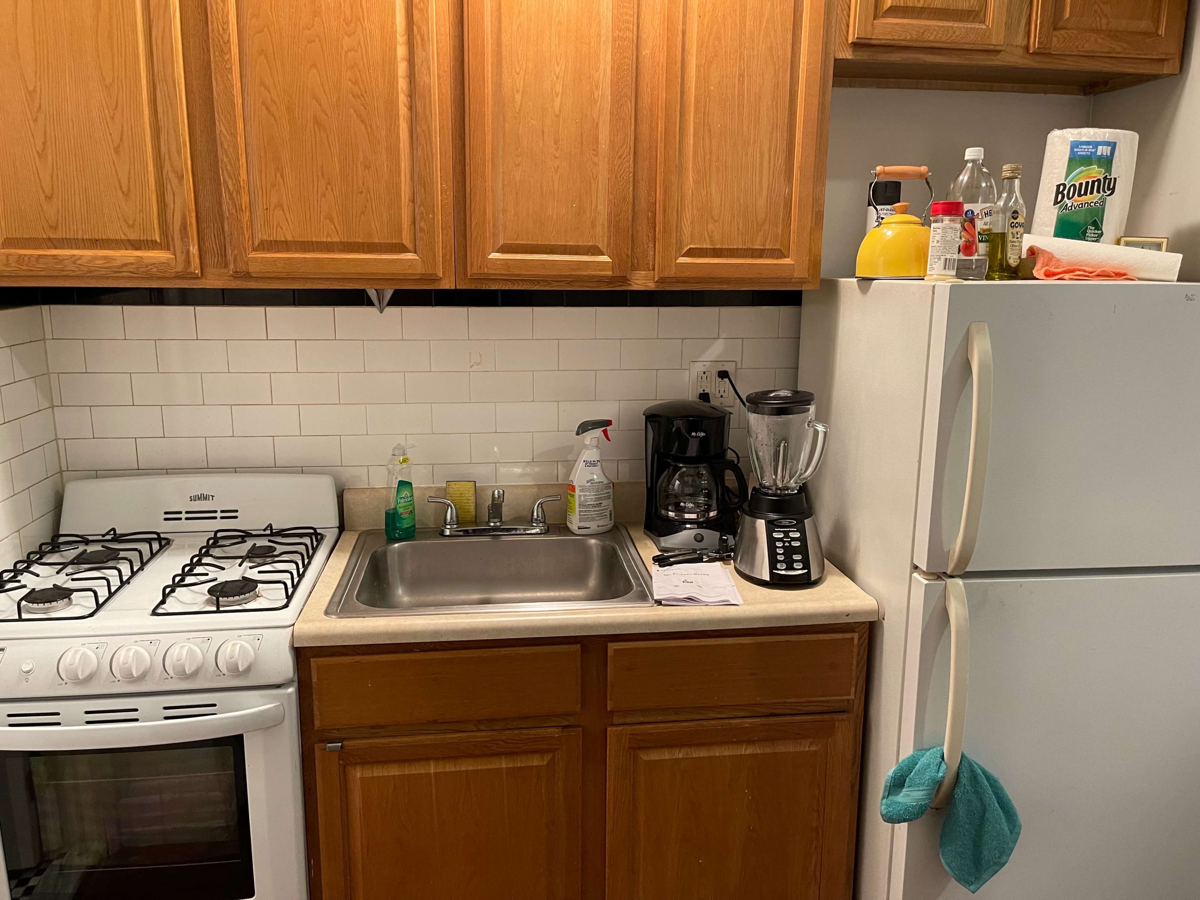 The image shows a kitchen area with wooden cabinets, a stainless steel sink, a gas stove, a blender, a coffee maker, and a refrigerator.