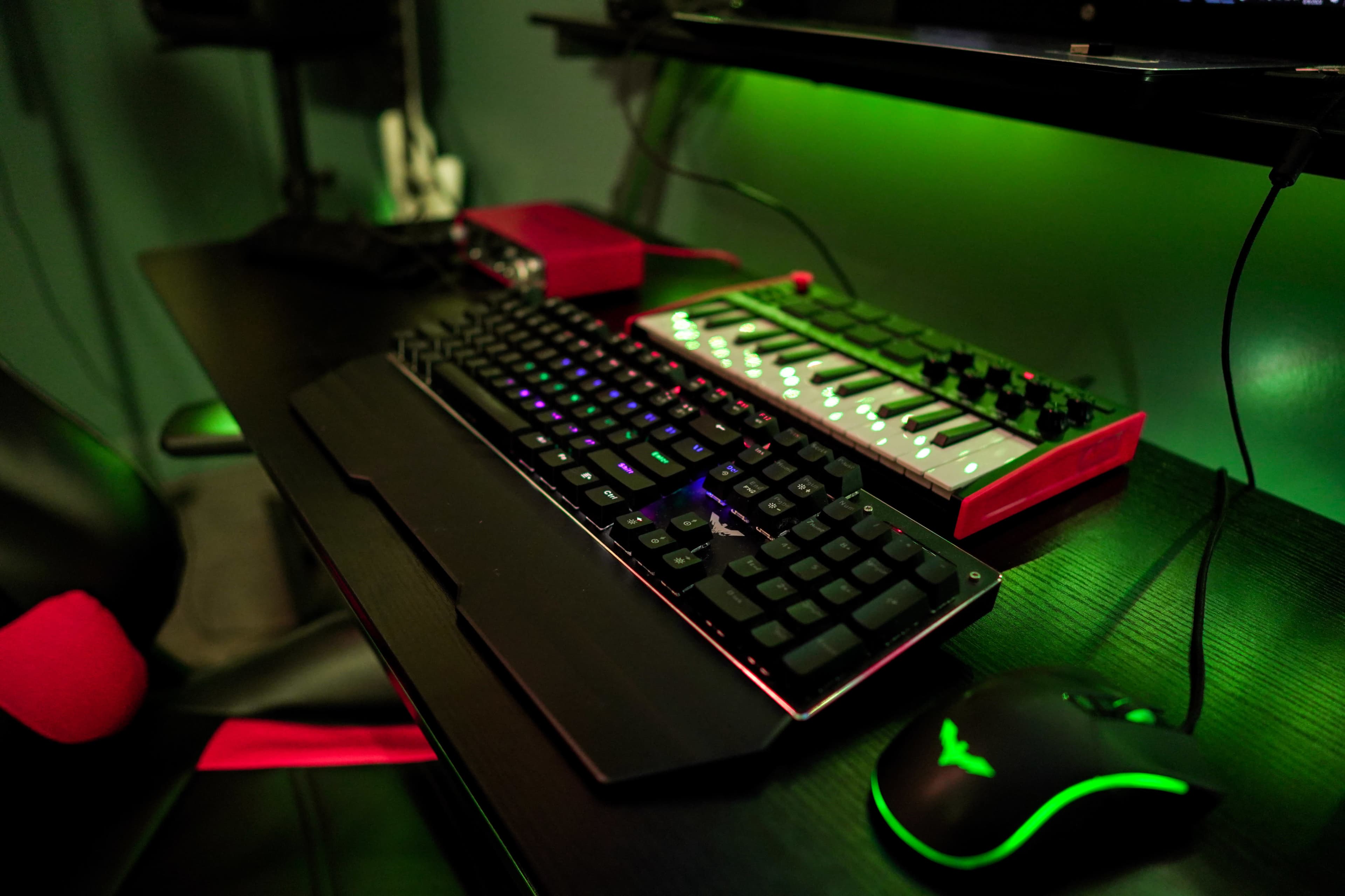 The image shows a gaming keyboard, a MIDI controller, and a computer mouse on a black desk with green lighting in the background.