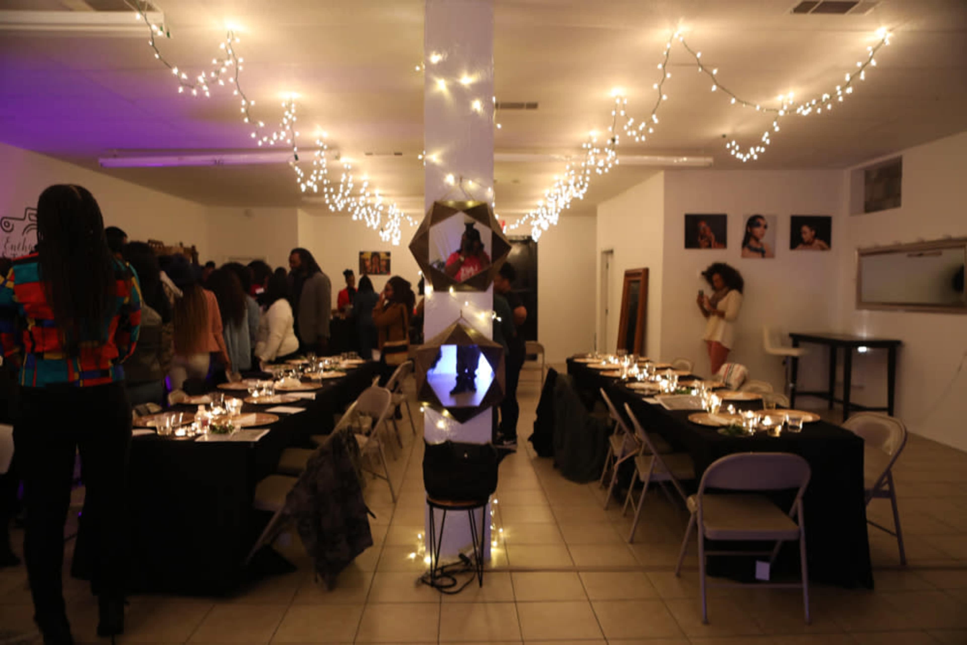 A festive gathering in a dimly lit room with tables set for dining, decorated with string lights and surrounded by guests mingling.