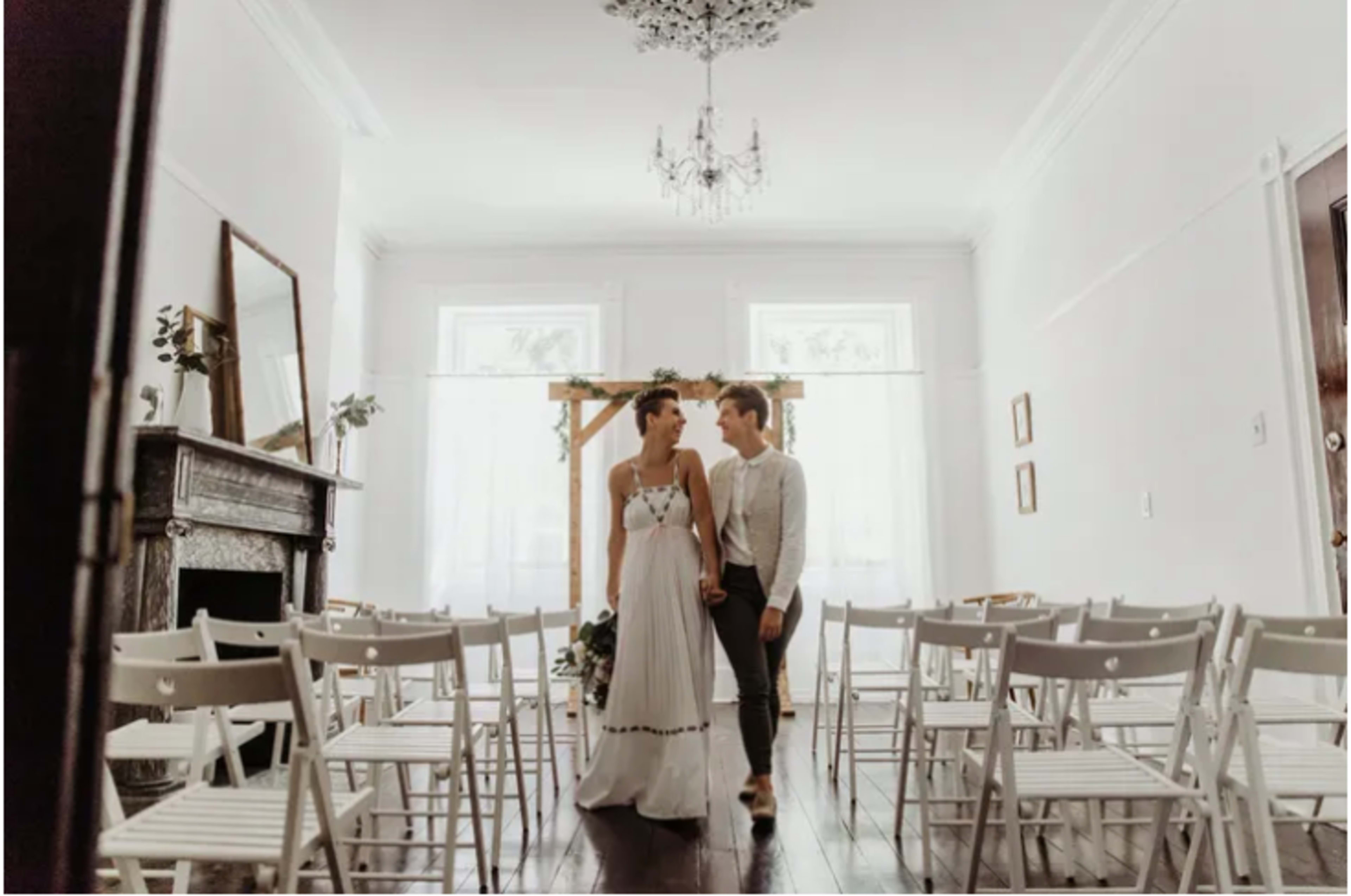 A bride and groom walk hand in hand down the aisle of a lightly decorated indoor venue set up for a wedding ceremony.