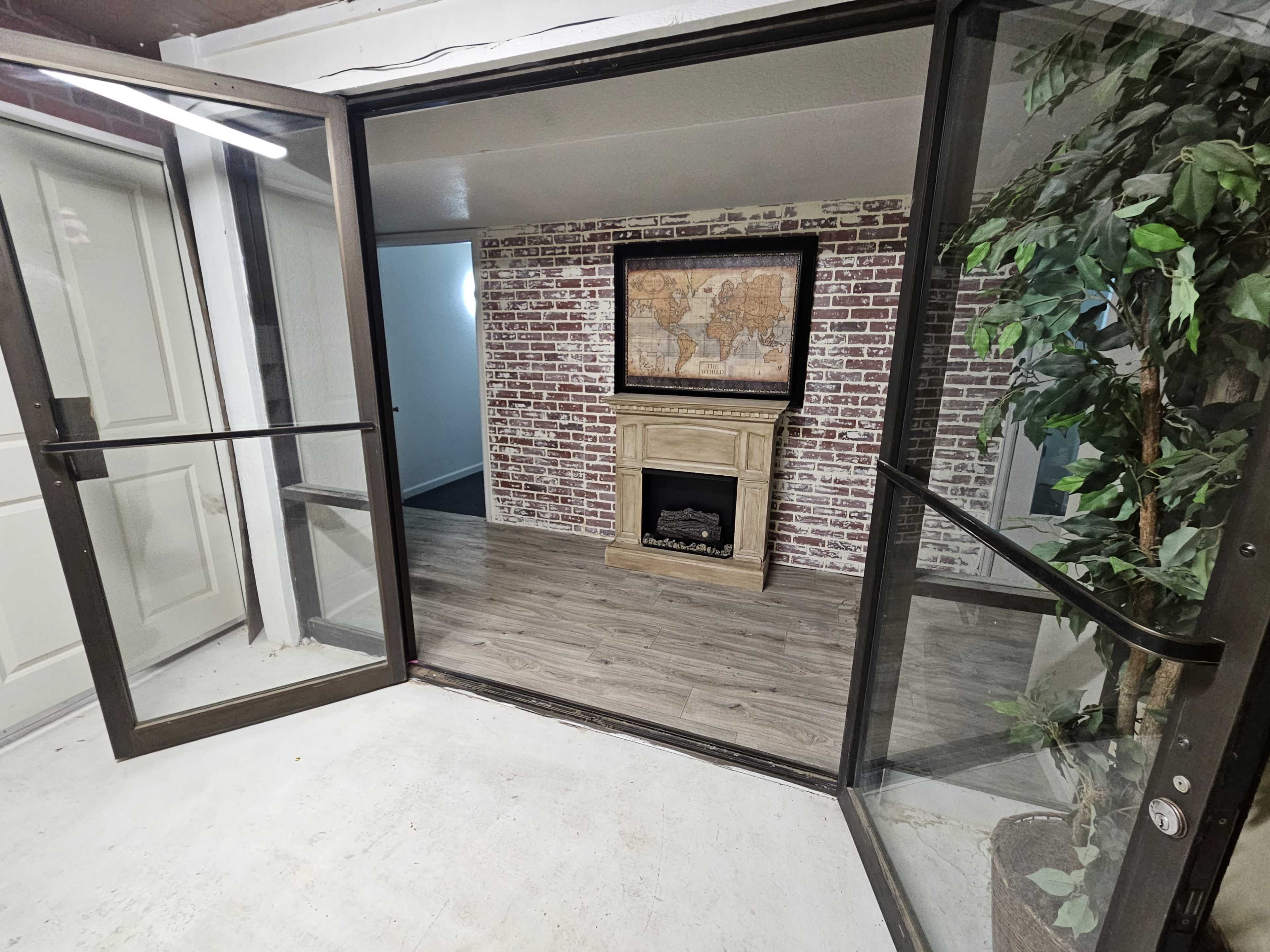 An enclosed room with glass doors leading to a space featuring a faux fireplace and a world map on a brick wall.