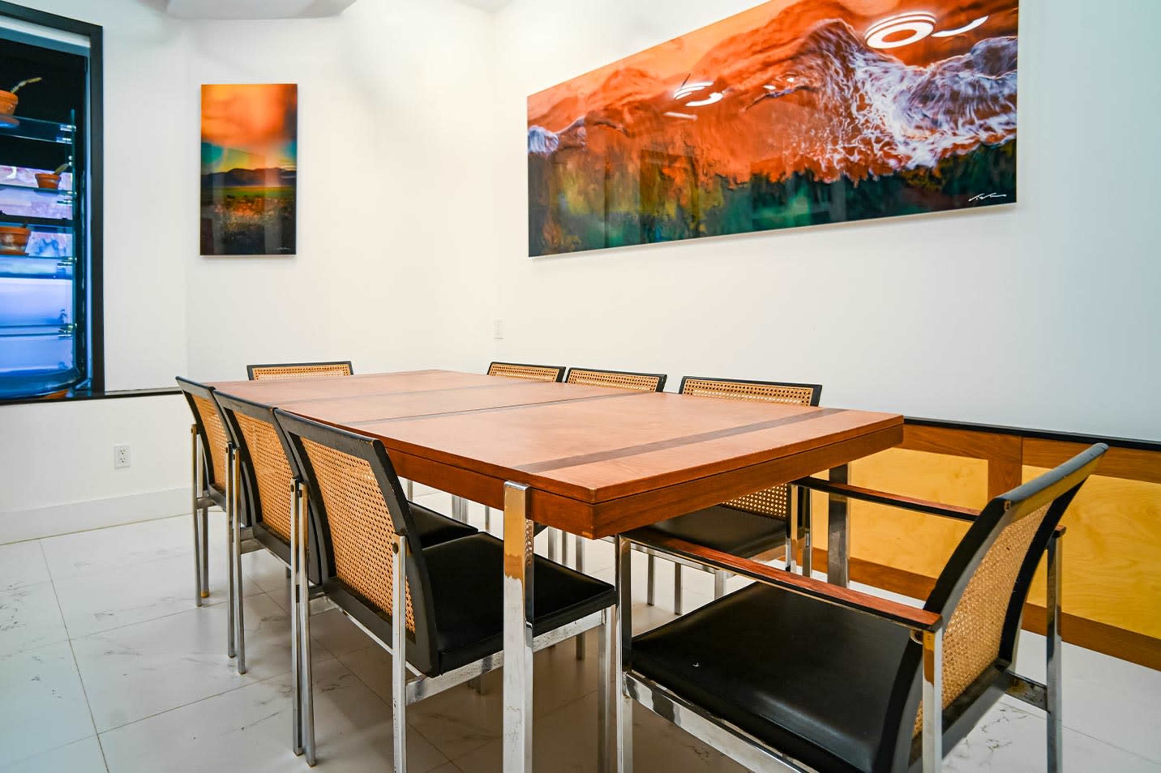 The image shows a modern dining area with a large wooden table surrounded by eight black chairs, and a piece of abstract art hanging on the wall.