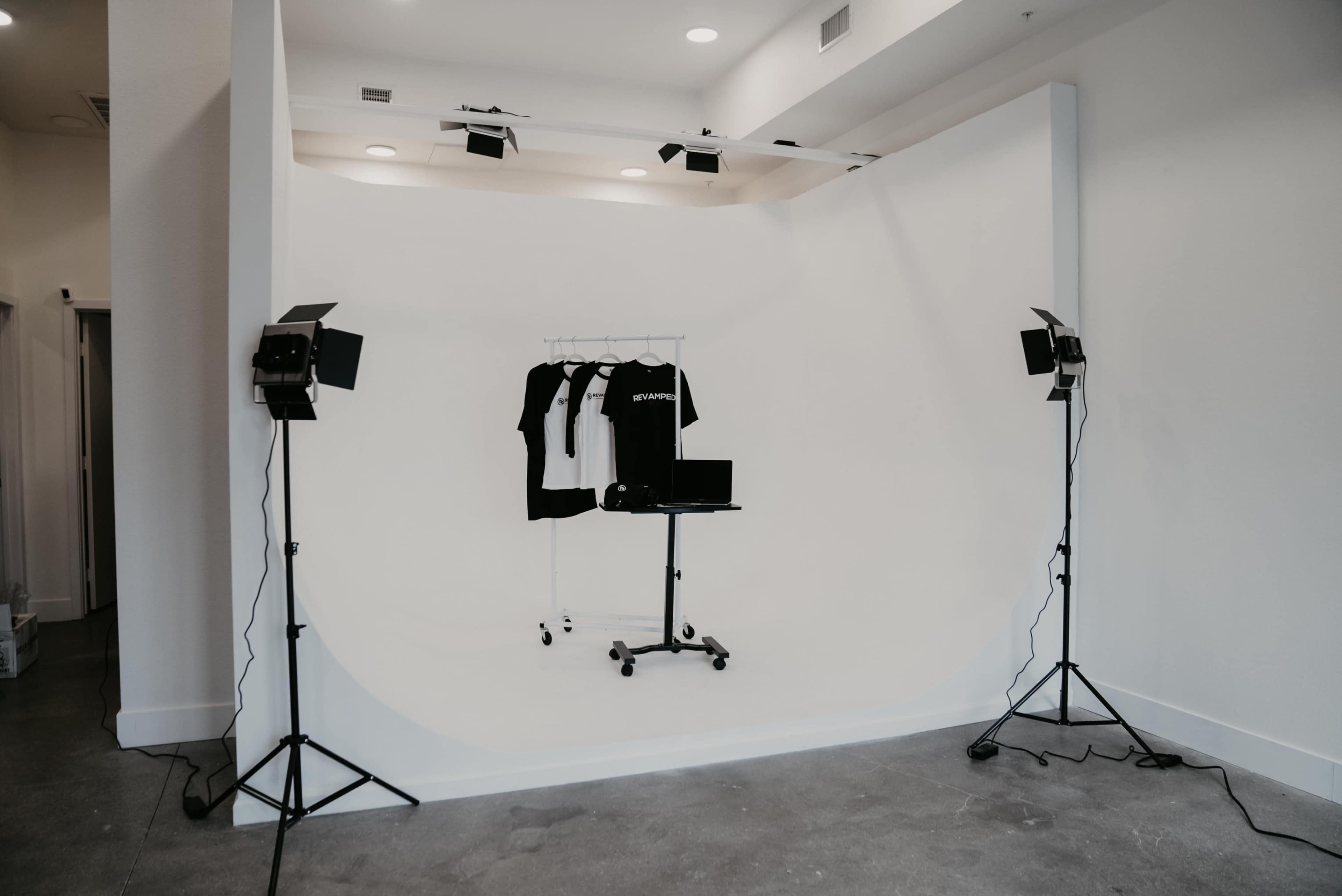A minimalist photography studio with a white backdrop, a clothing rack holding black and white shirts, and lighting equipment arranged on either side.