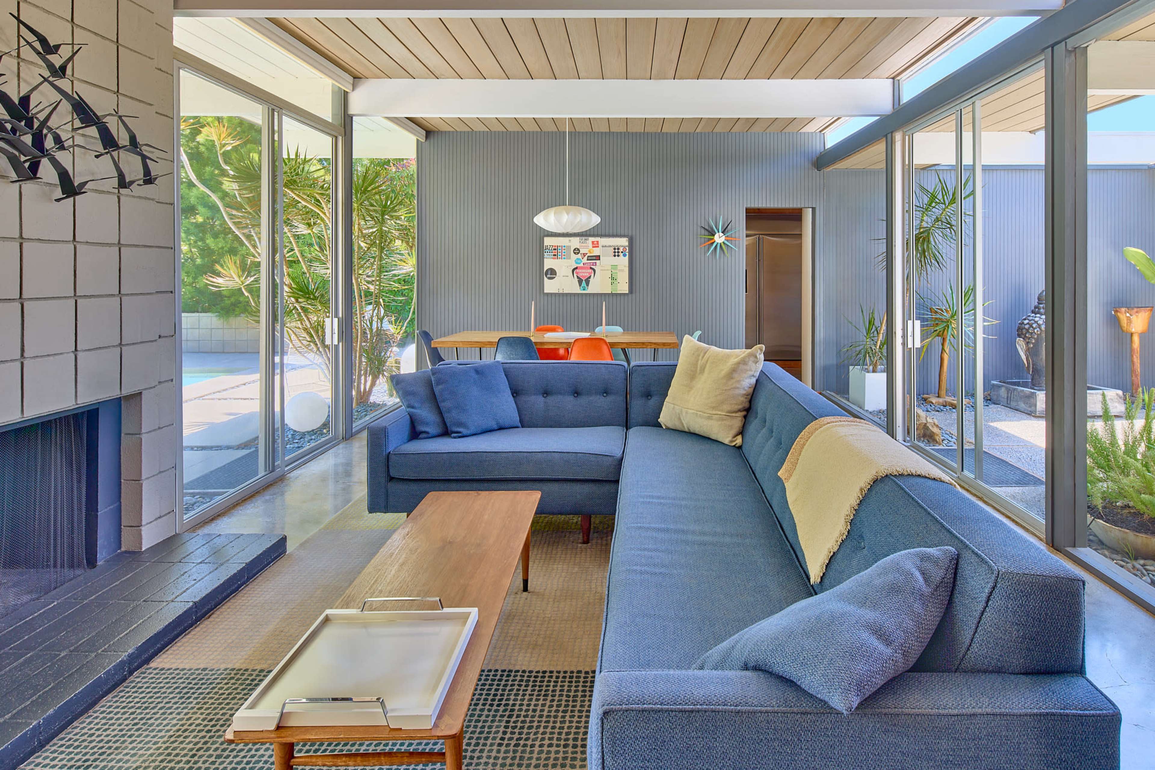 The image shows a modern living room with a sectional couch, a coffee table, and large glass windows that provide a view of an outdoor space with plants.
