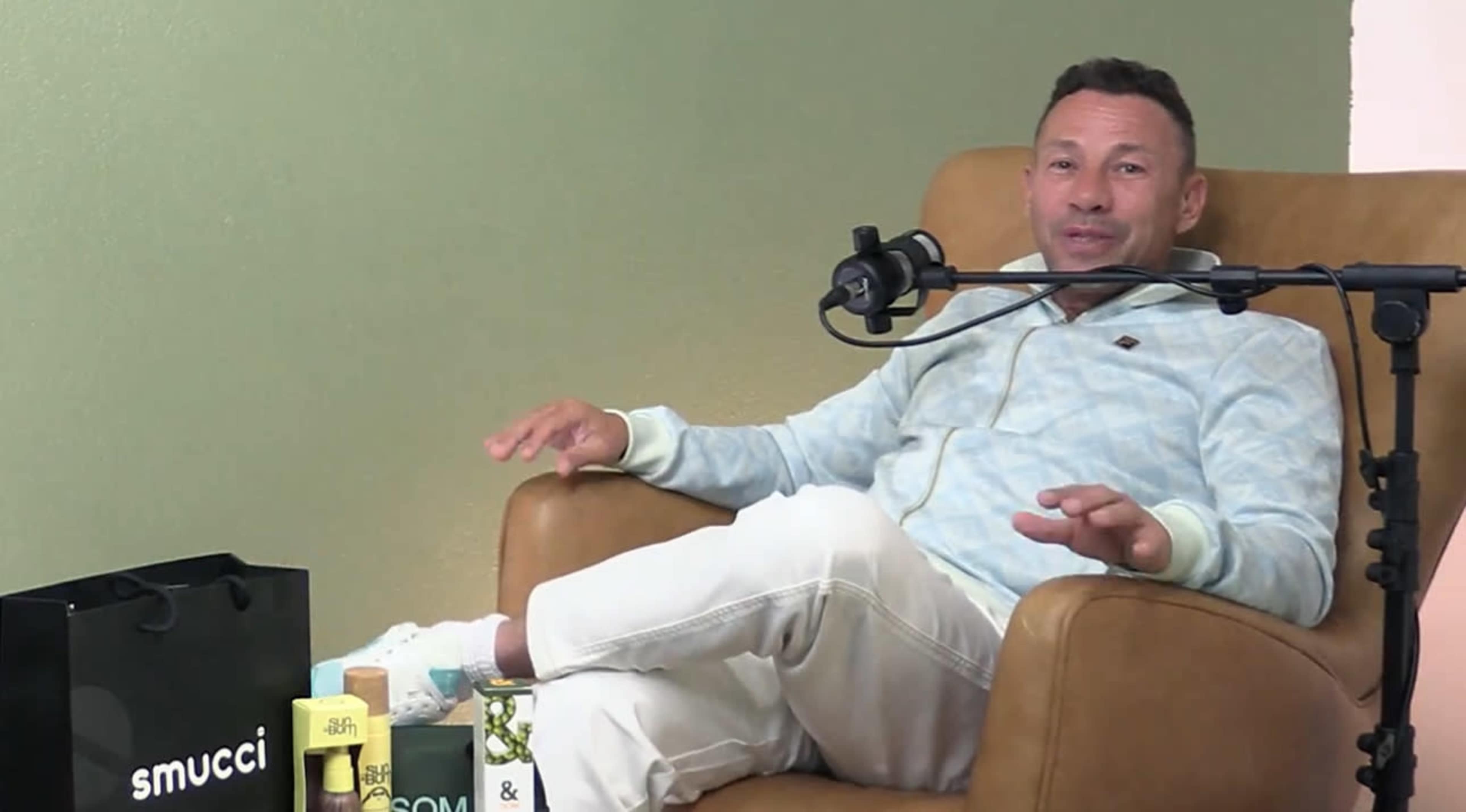 A man is sitting in a brown chair, speaking into a microphone with various products placed on a table beside him.