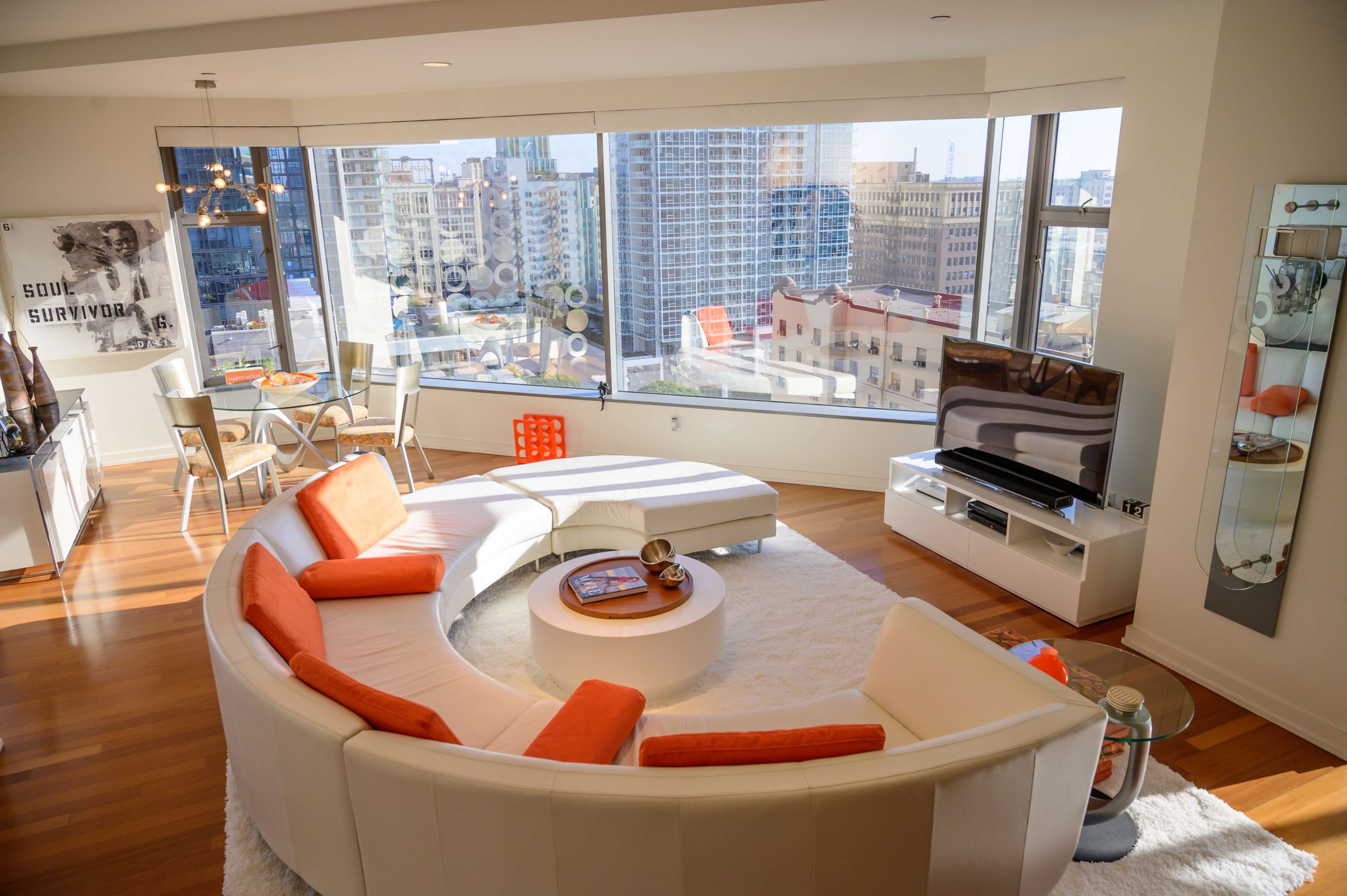 A modern living room with a circular white sofa, orange accents, a television, and a large window offering a city view.