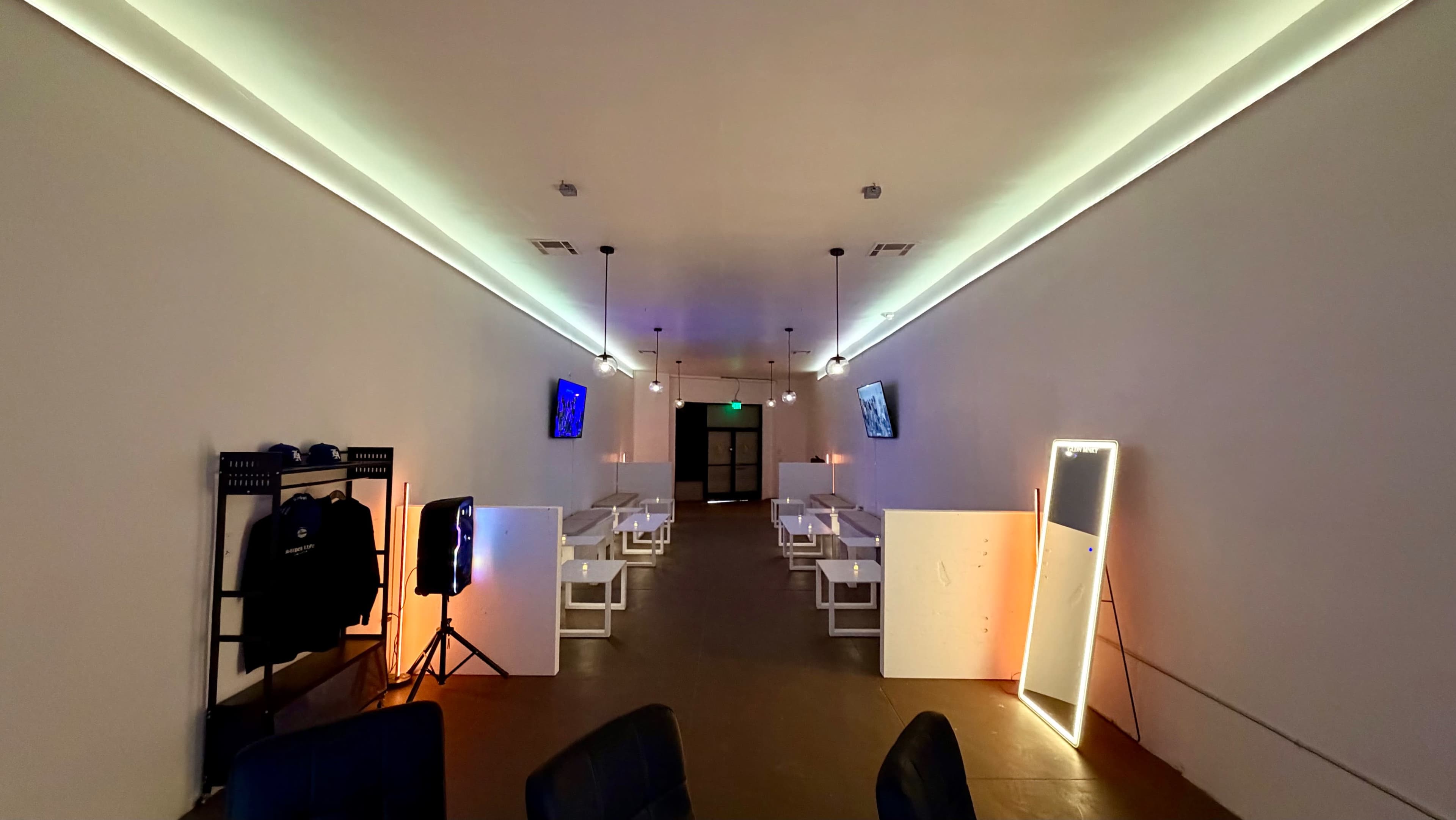 The interior of a modern, minimalist venue features white walls, sleek seating arrangements, and ambient lighting with two screens mounted on the walls.