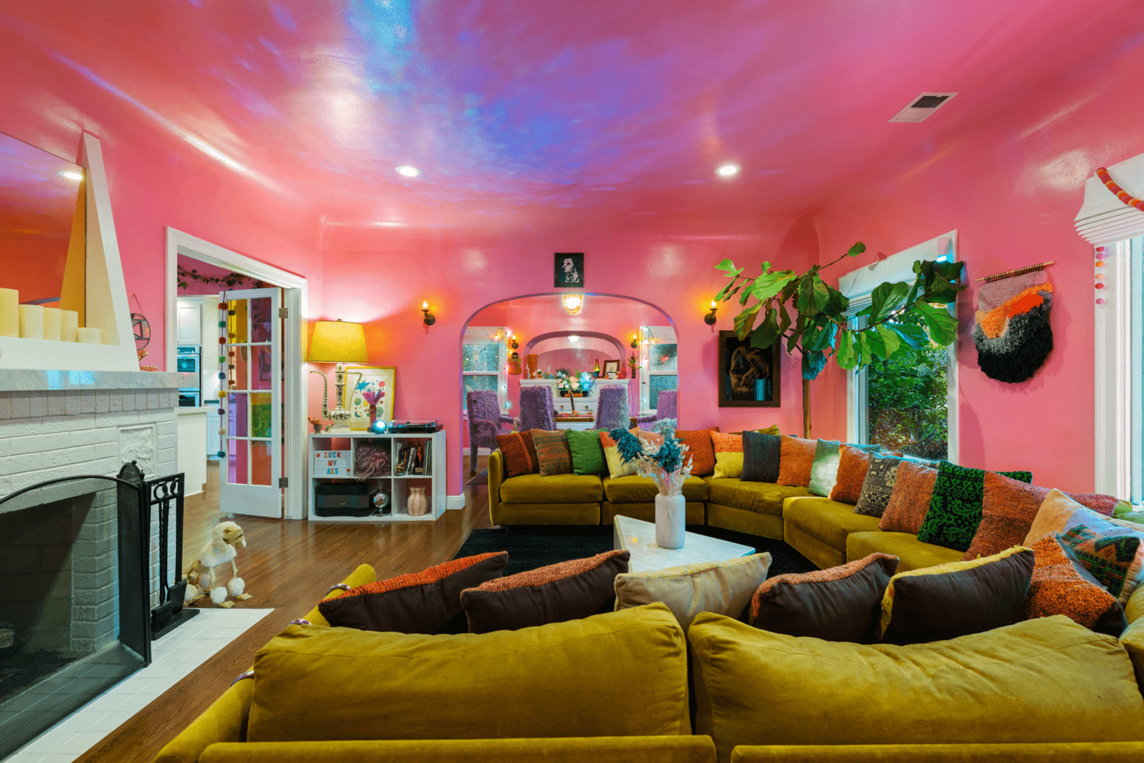 A bright pink living room with a large yellow sectional sofa, a marble coffee table, and various decorative elements, including plants and colorful throw pillows.