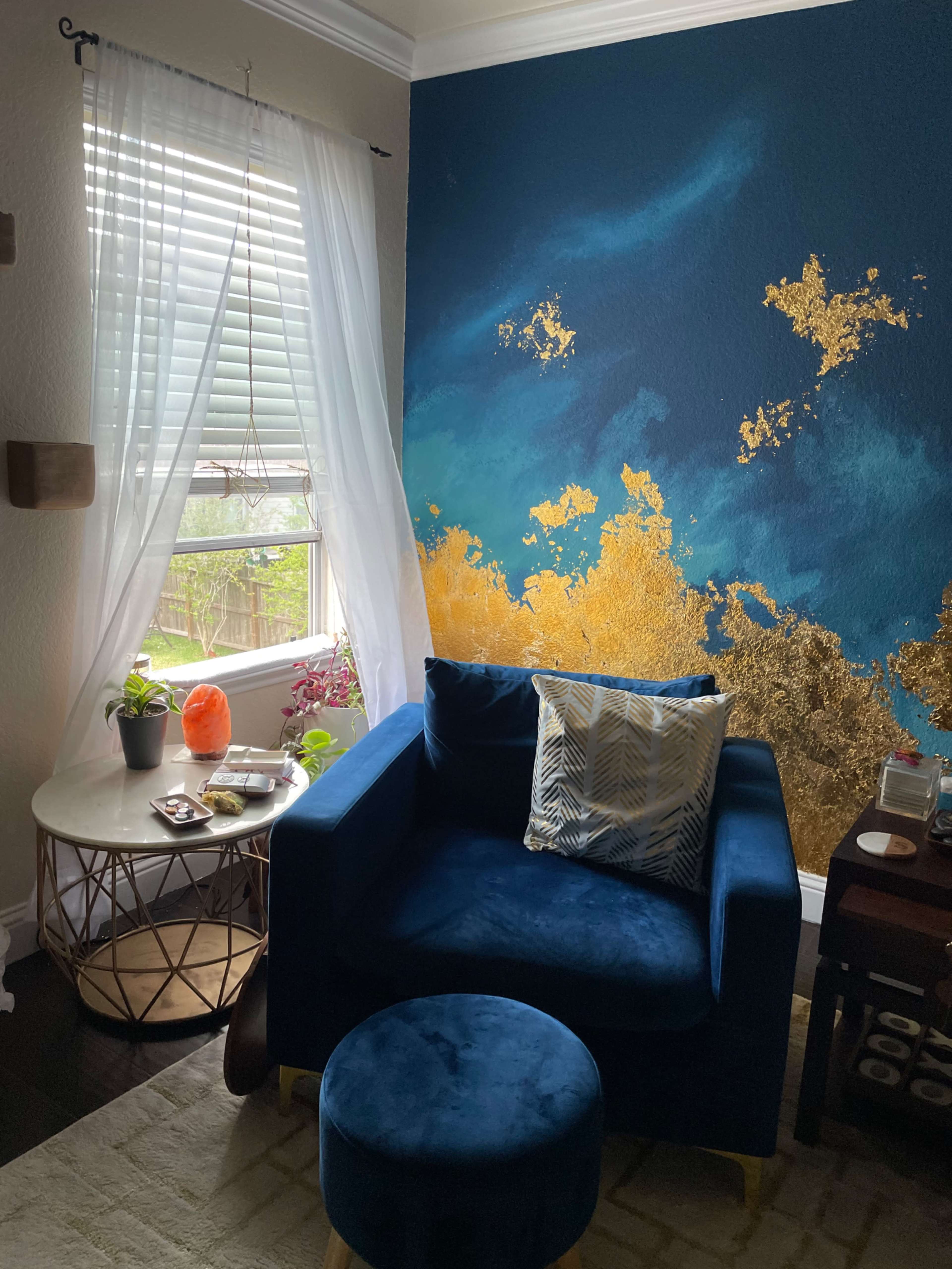 A blue velvet armchair and matching ottoman are positioned beside a round table adorned with decorative items and plants, with a large window letting in natural light.