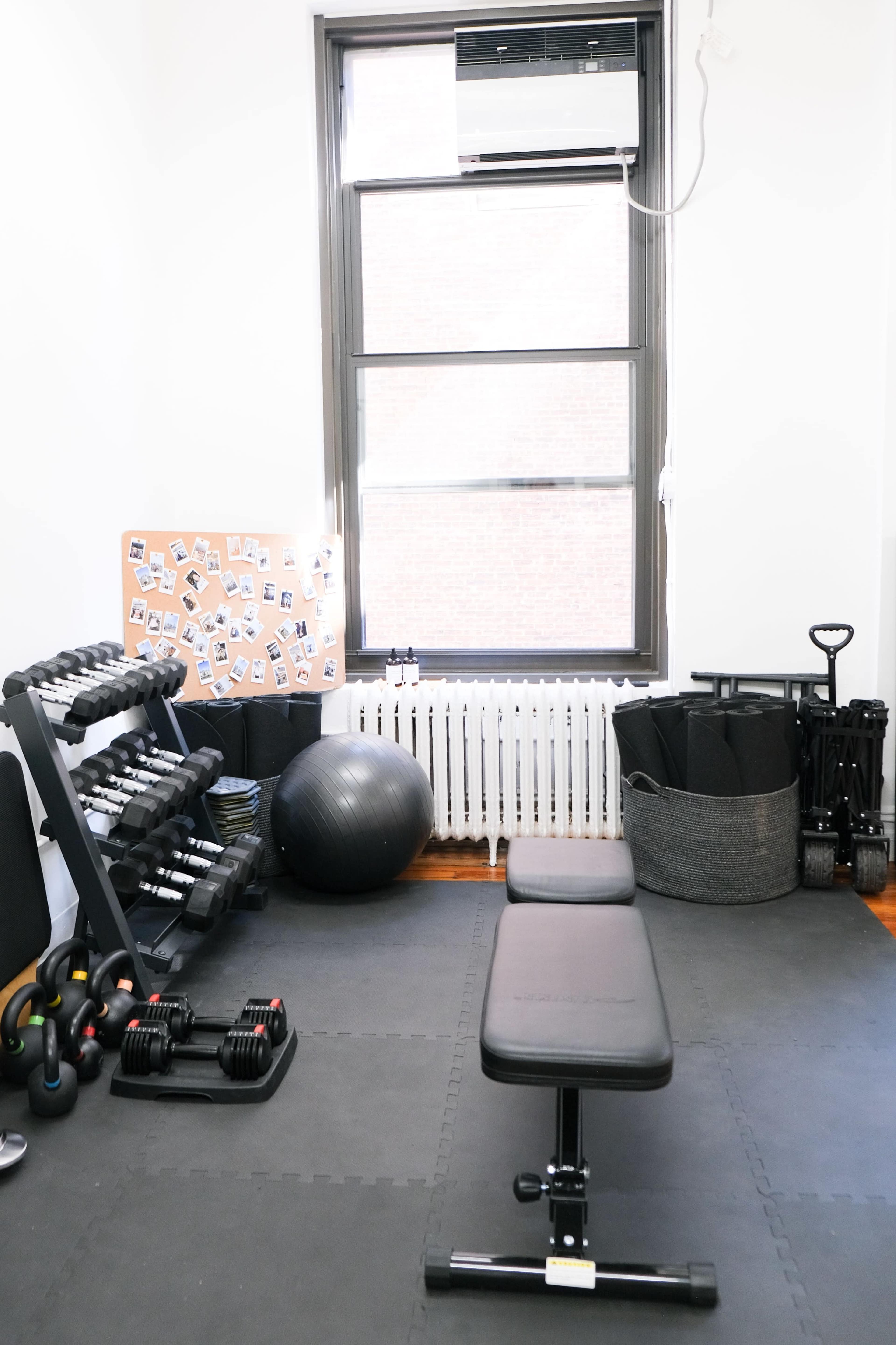 The image shows a home gym set up with dumbbells on a rack, a black exercise bench in the center, a stability ball, and a storage bin against a window.
