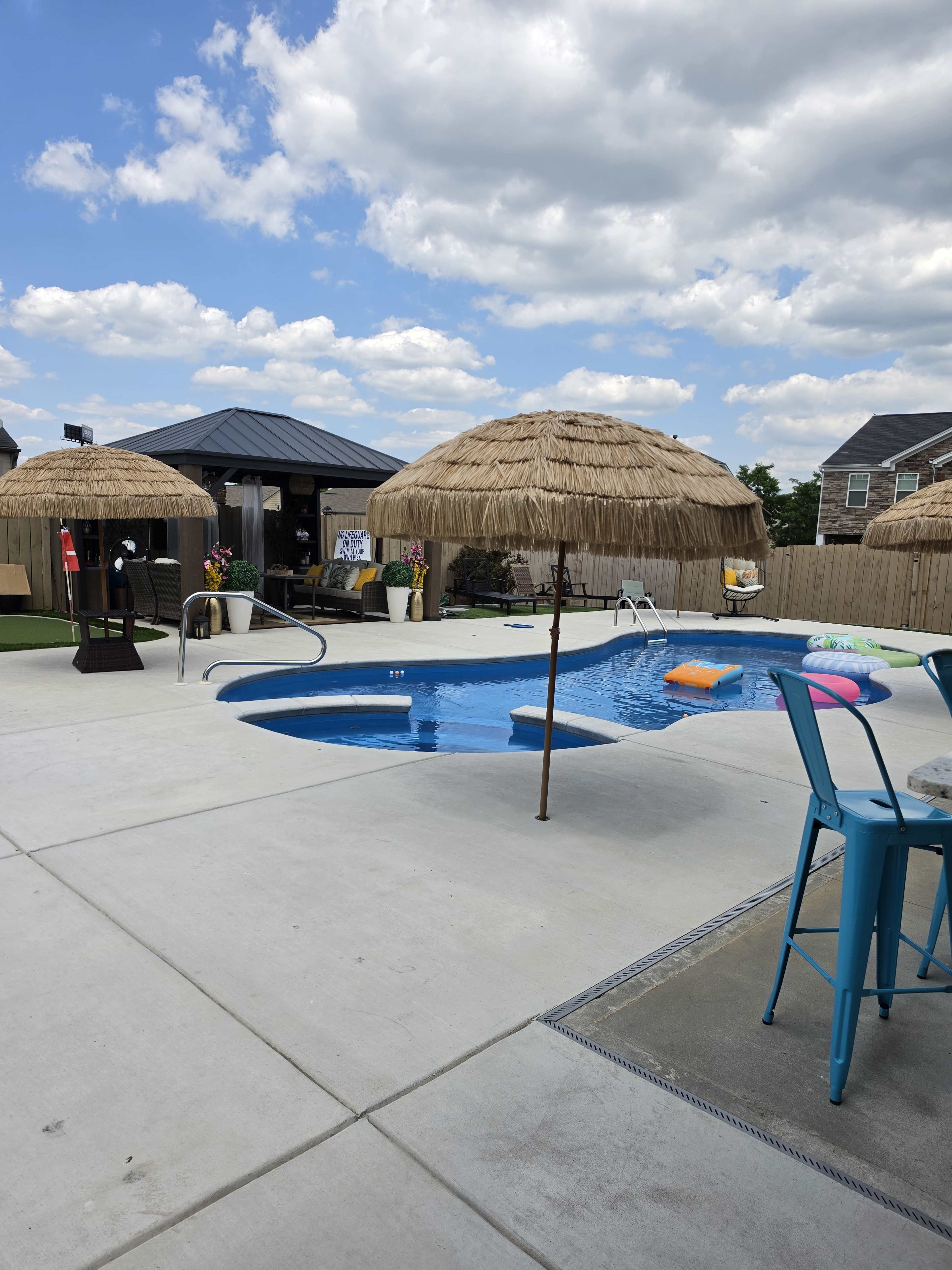 A swimming pool with thatched umbrellas and surrounding lounge furniture is situated in a backyard area.